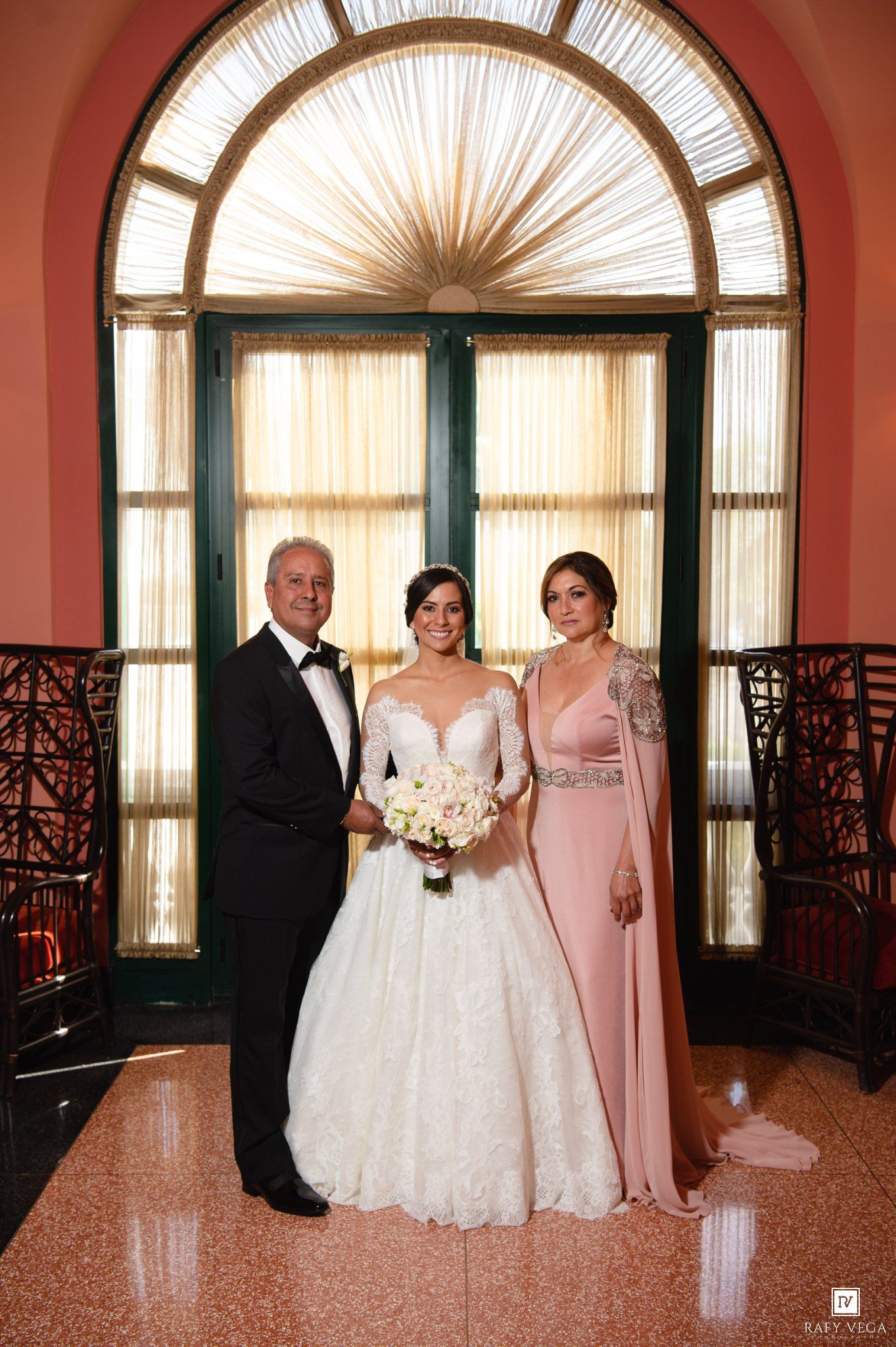 Bride in white gown poses with parents in elegant pink venue, holding bouquet before arched window.