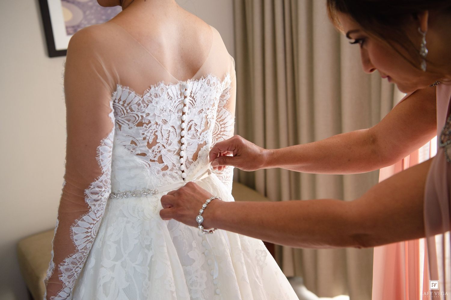 Bridesmaid fastening buttons on back of elegant lace wedding dress during bridal preparation.