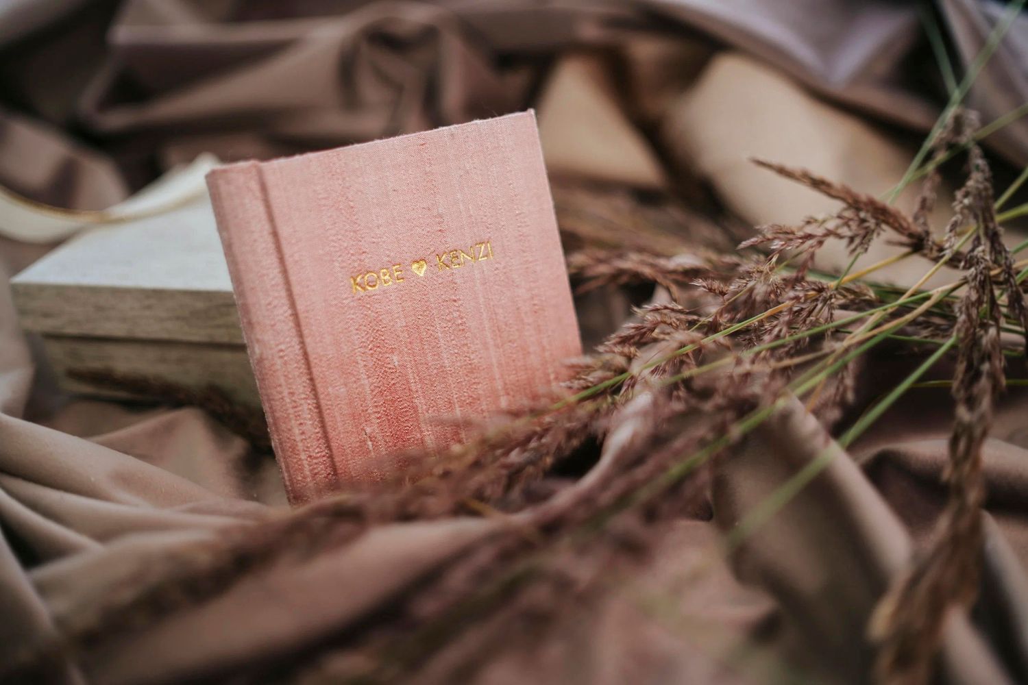 A pink hardcover book with gold lettering rests among dried grass stems on soft brown fabric.