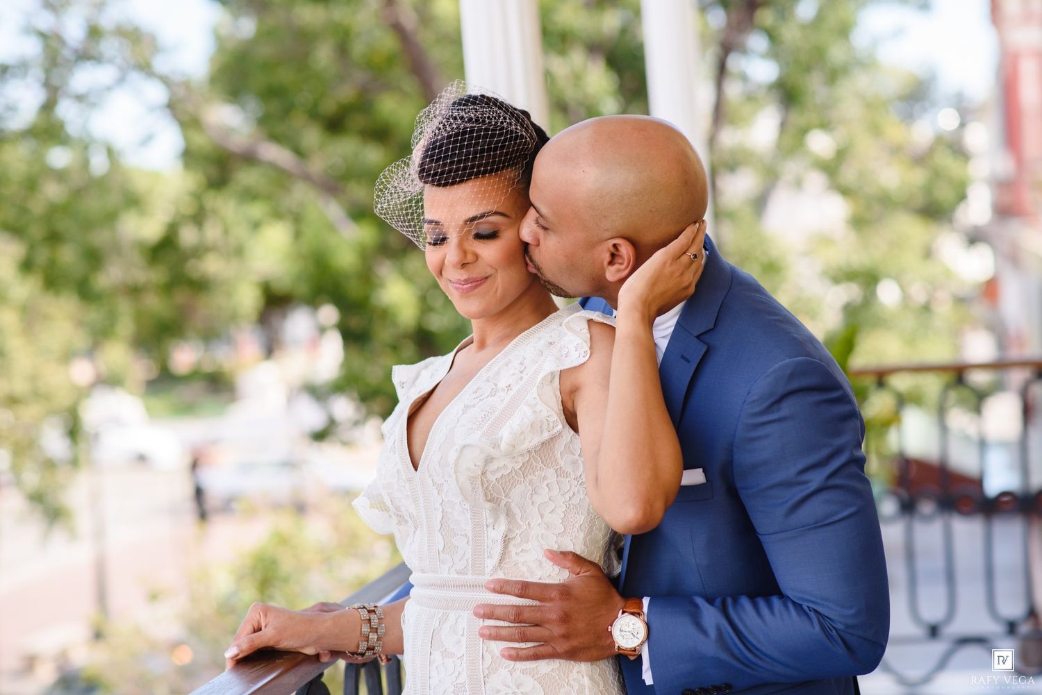 A groom in a blue suit kisses his bride wearing a white lace dress and birdcage veil on an outdoor balcony.