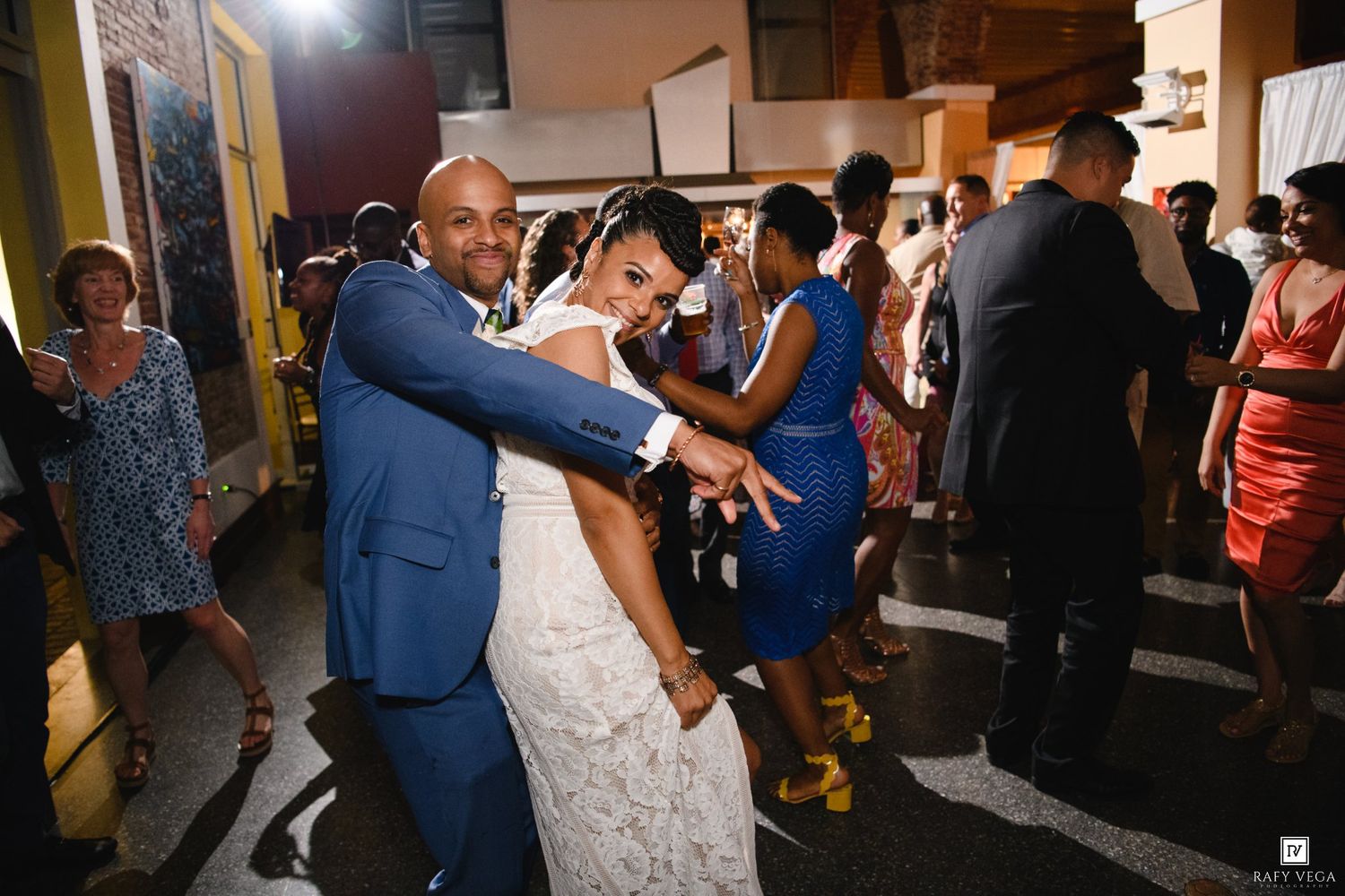 Bride and groom dancing joyfully at wedding reception, guests celebrating in background, groom in blue suit.