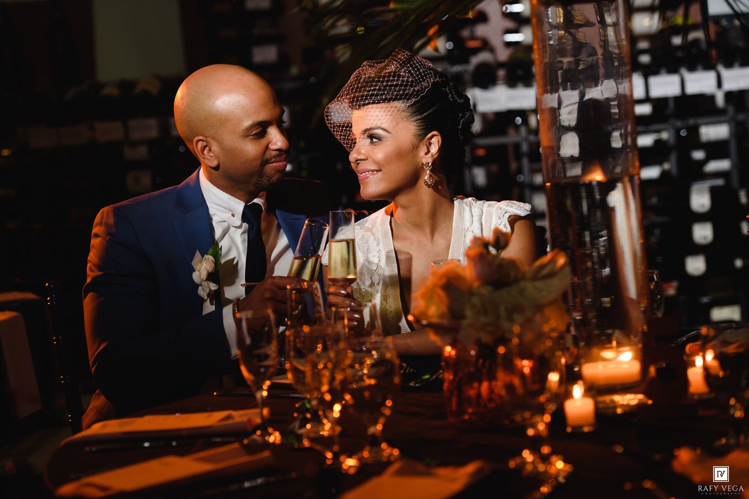 Couple in wedding attire sharing a romantic candlelit dinner, groom in blue suit, bride in white dress with fascinator.