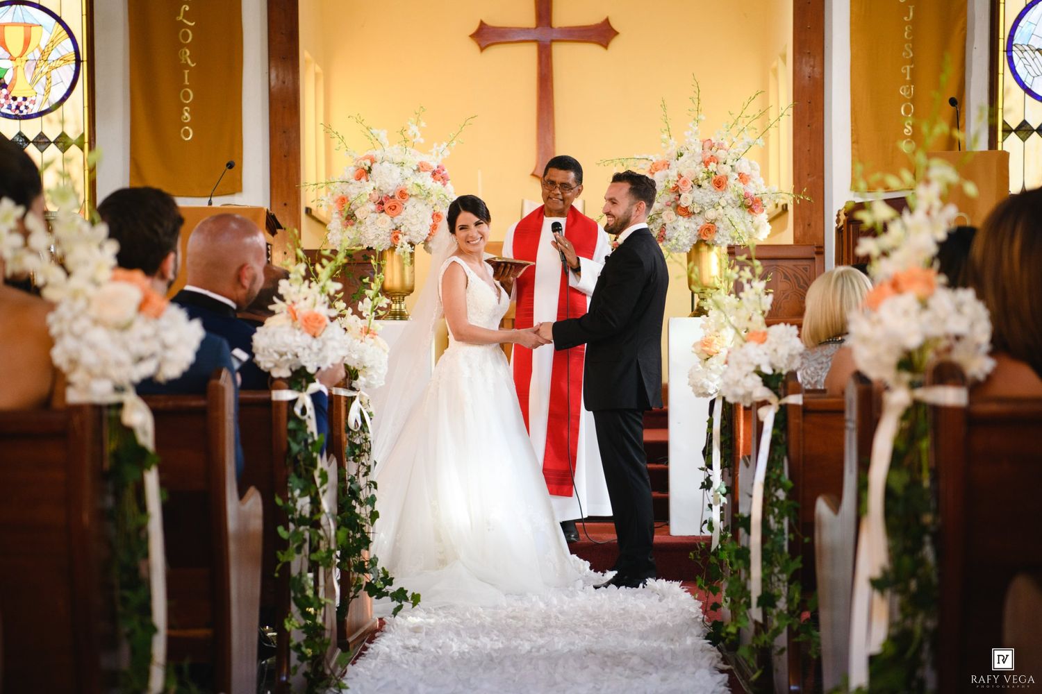 Couple exchanging vows at a church wedding ceremony with floral arrangements and a priest in red vestments.
