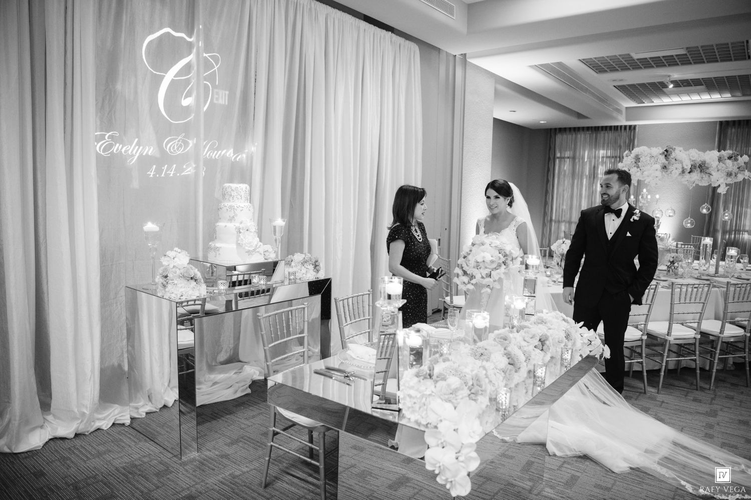 Black and white photo of bride and groom entering elegant wedding reception hall with floral decorations and cake display.