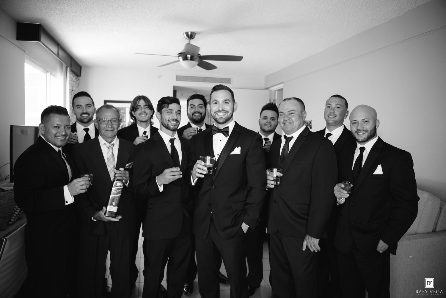 Groom and groomsmen in black tuxedos posing with drinks in black and white group wedding photo.