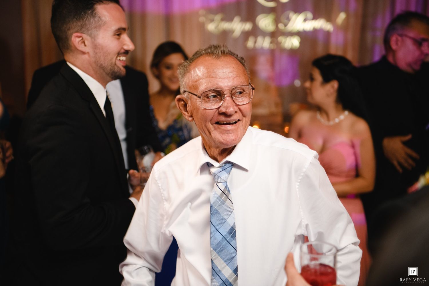 Elderly man smiling at a wedding reception, wearing a white shirt and blue tie, with guests in the background.