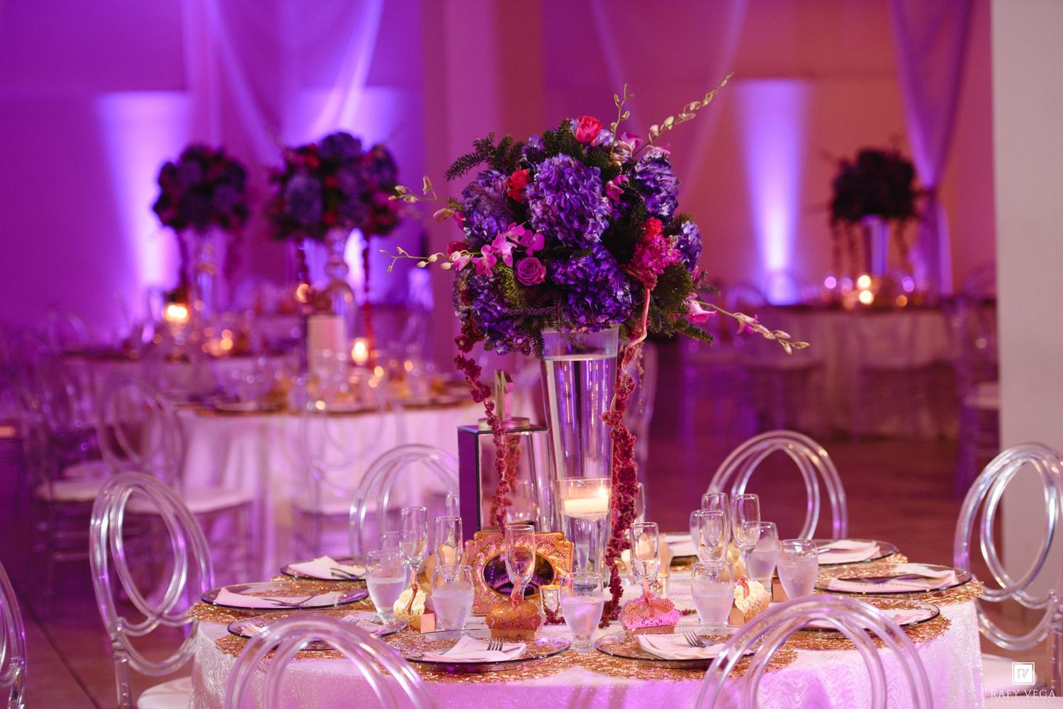 Elegant wedding reception with purple lighting, ghost chairs, floral centerpieces, and candlelit round tables.