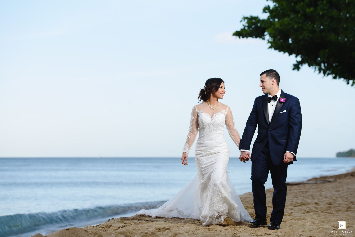 Bride and groom walk hand-in-hand on a sandy beach, she in a lace gown, he in a navy suit.