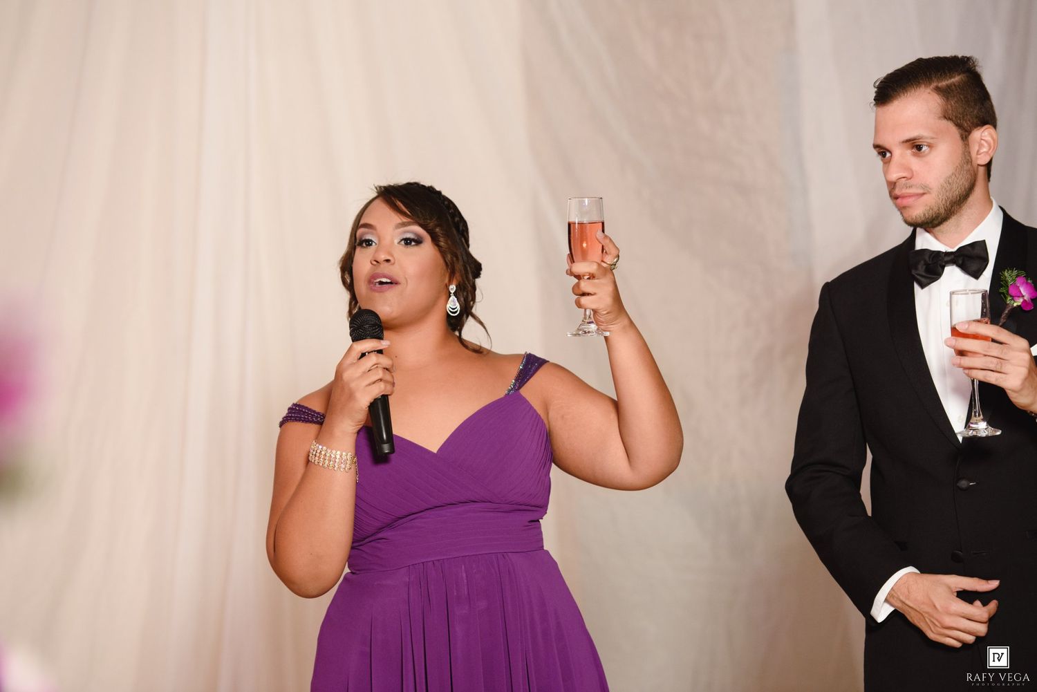 Bridesmaid in purple dress giving a toast at a wedding reception while a groomsman in a tuxedo stands nearby.