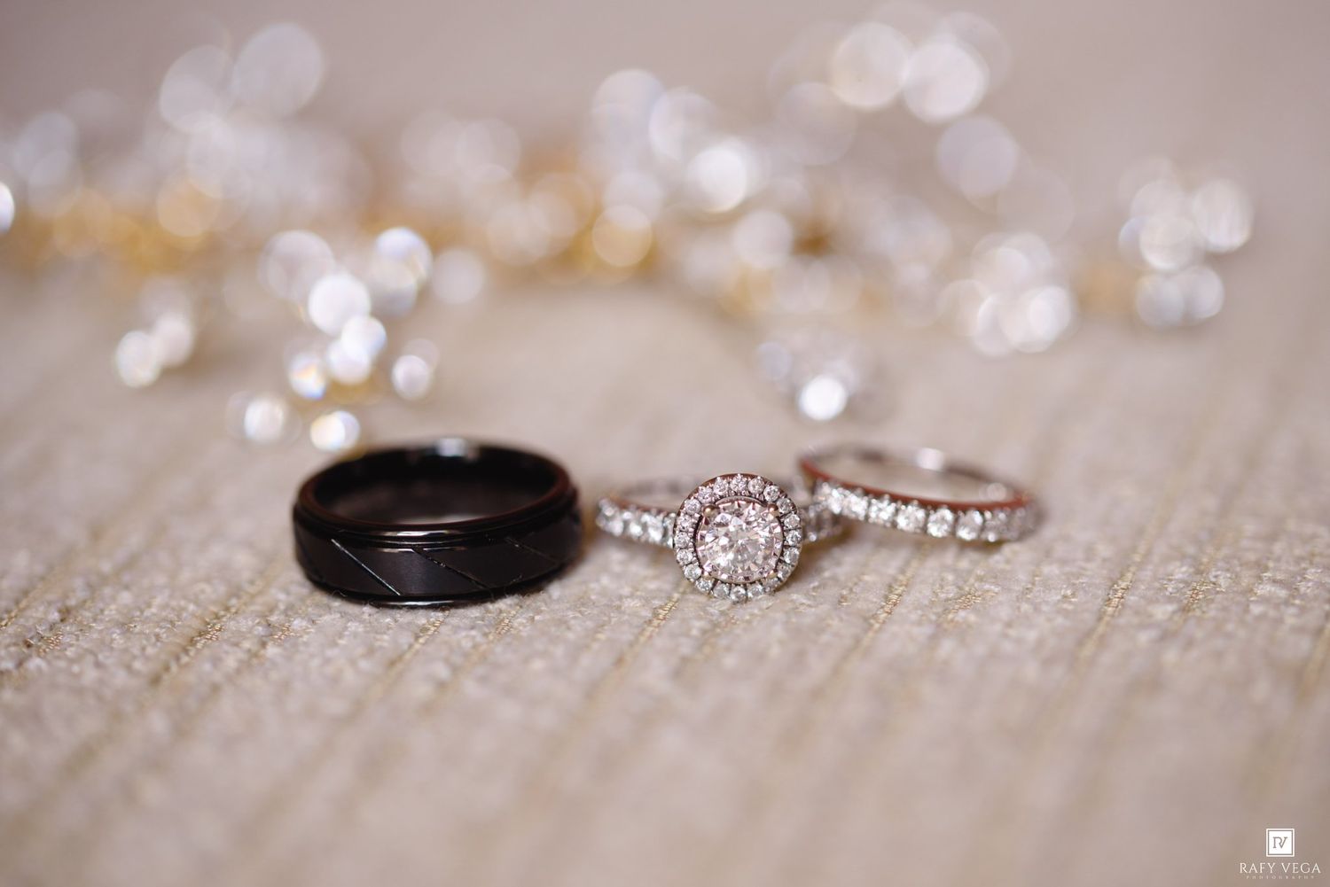 Three wedding rings displayed on textured fabric, featuring a black band, diamond halo engagement ring, and diamond wedding band.