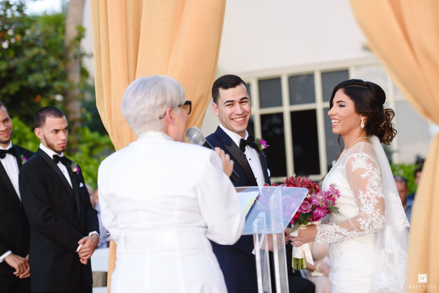 Outdoor wedding ceremony with bride and groom exchanging vows before an officiant under golden drapes.