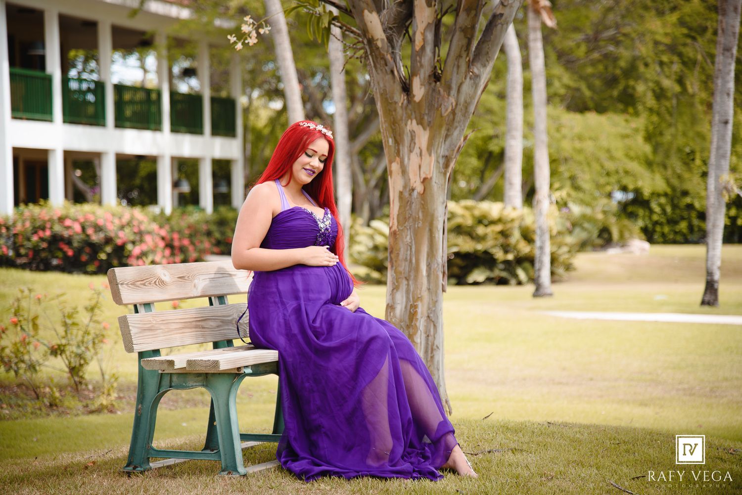 Pregnant woman in purple maternity gown sitting on bench under tree in lush green park setting.