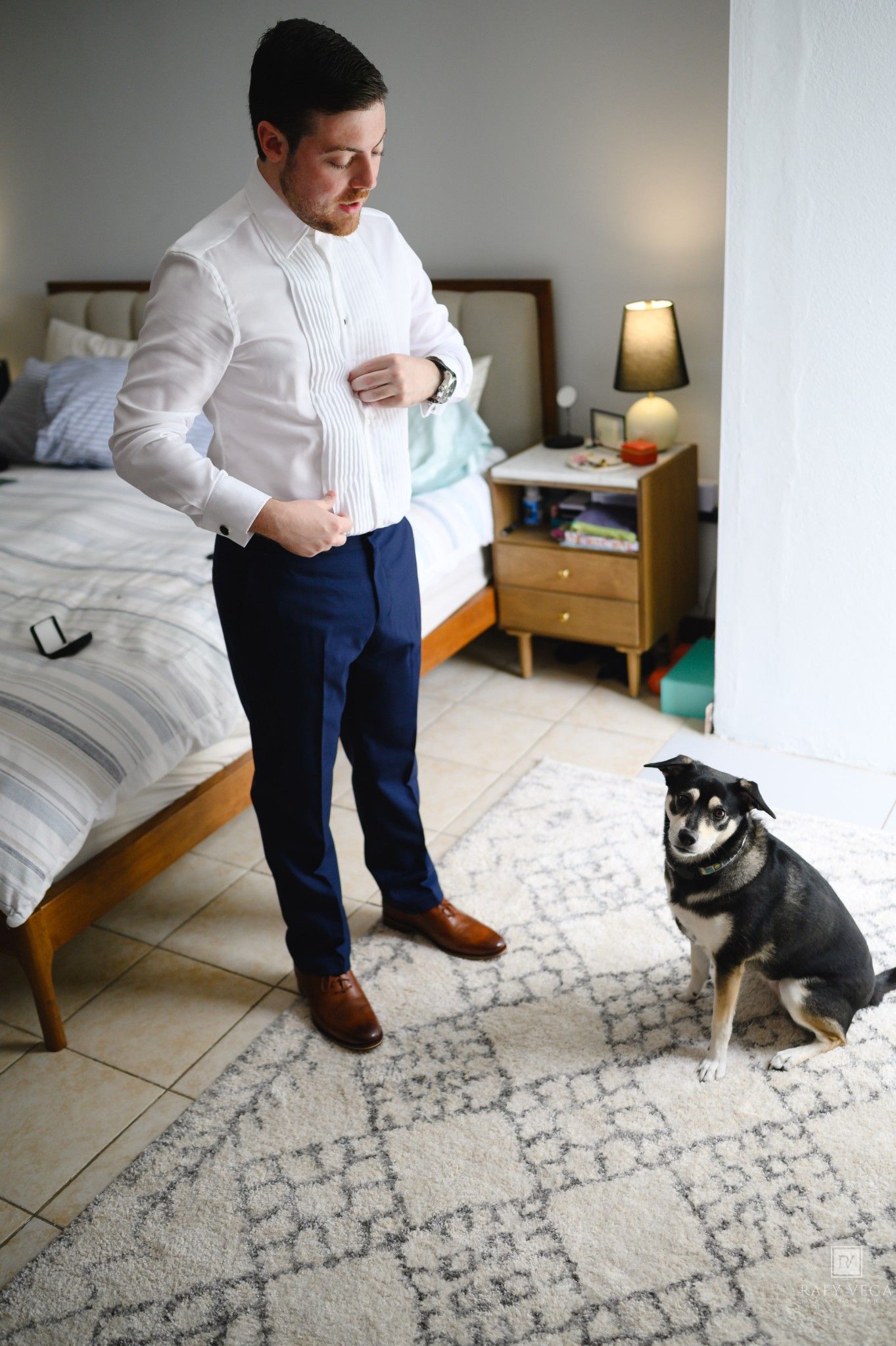 Man in white shirt and blue trousers buttoning up while small black dog sits on rug in bedroom.