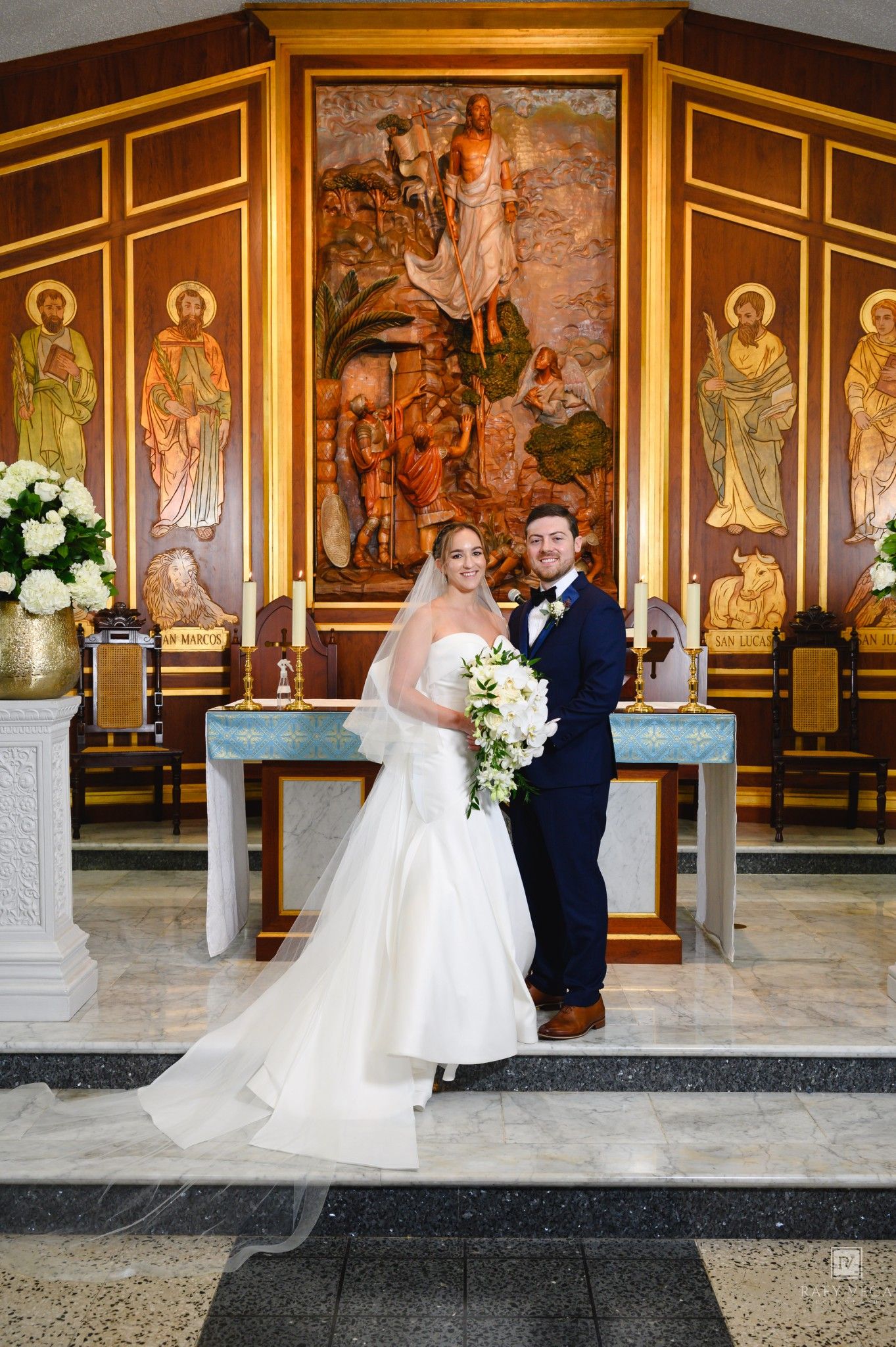 Bride and groom posing at church altar with ornate religious murals, bride in mermaid gown holding white bouquet.