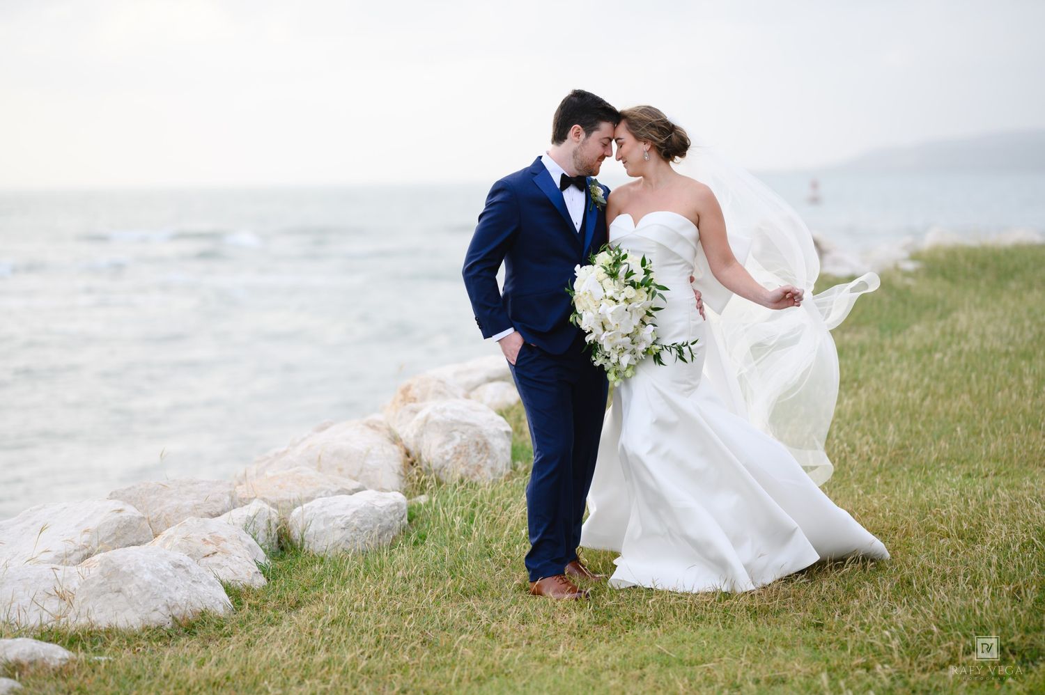 Bride and groom share a romantic moment by the seaside, her white gown flowing in the wind, holding a white bouquet.