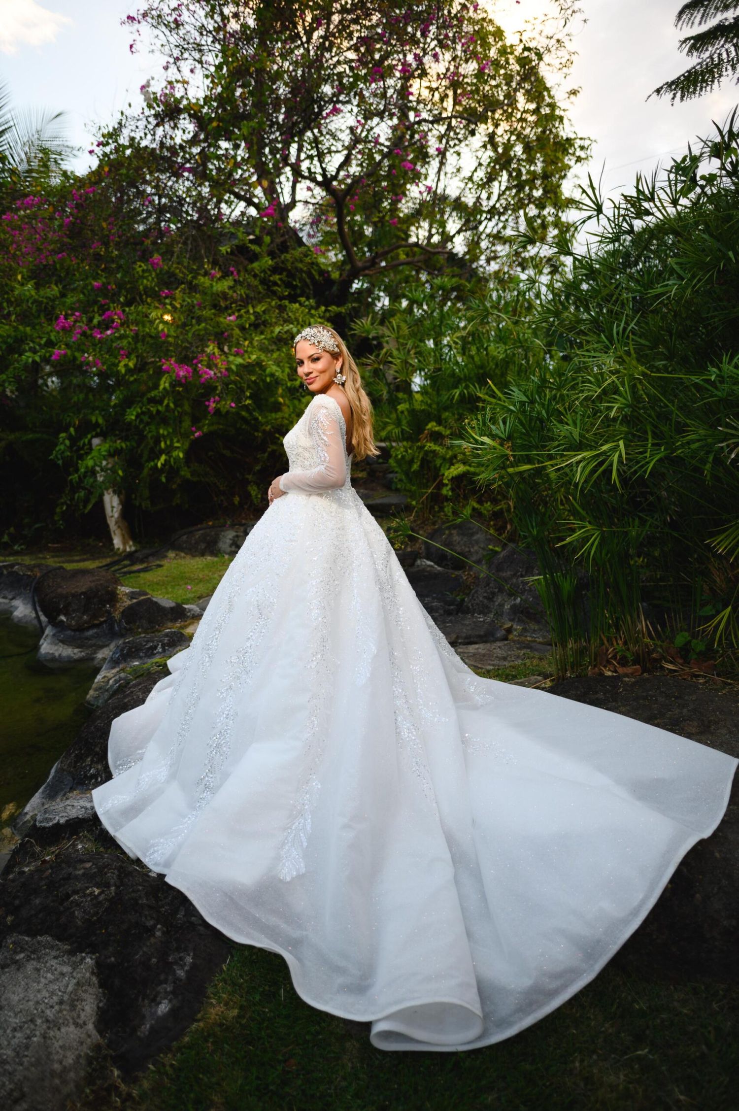 Bride in elegant long-sleeve lace ball gown with cathedral train poses in lush tropical garden setting.