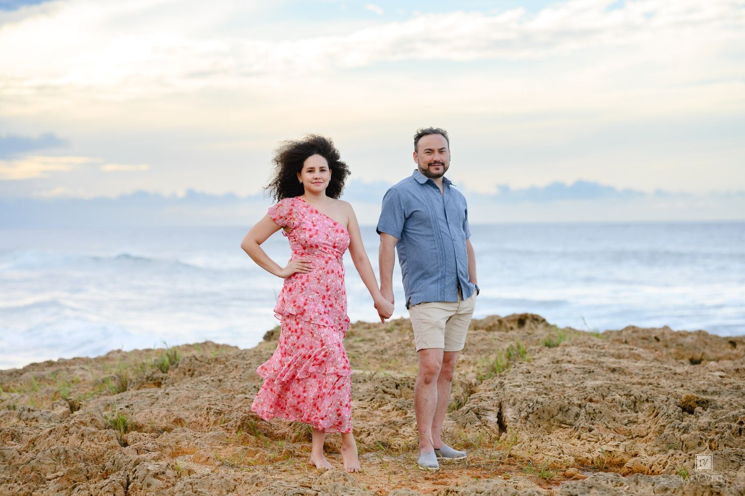Couple holding hands on rocky coastal cliffs with ocean waves and cloudy sky backdrop.