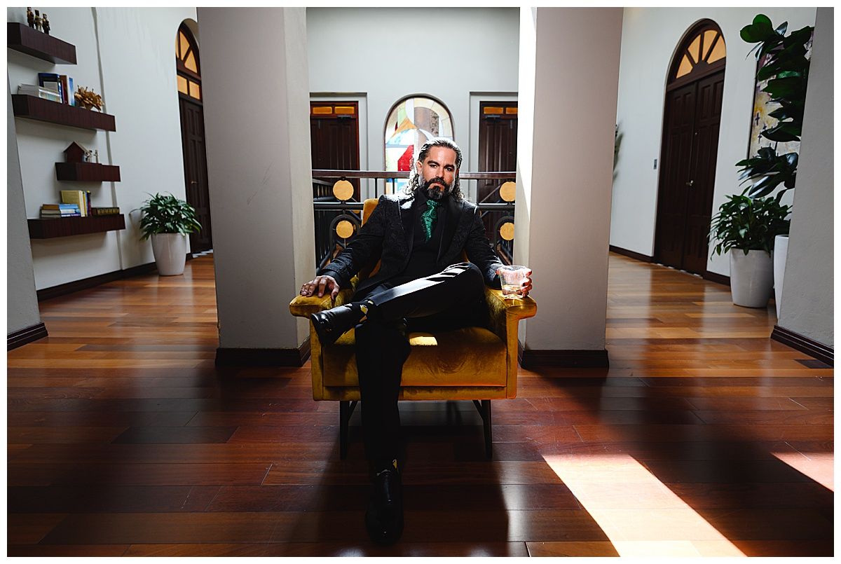 Man in suit sitting confidently in a yellow armchair in an elegant hallway with hardwood floors.