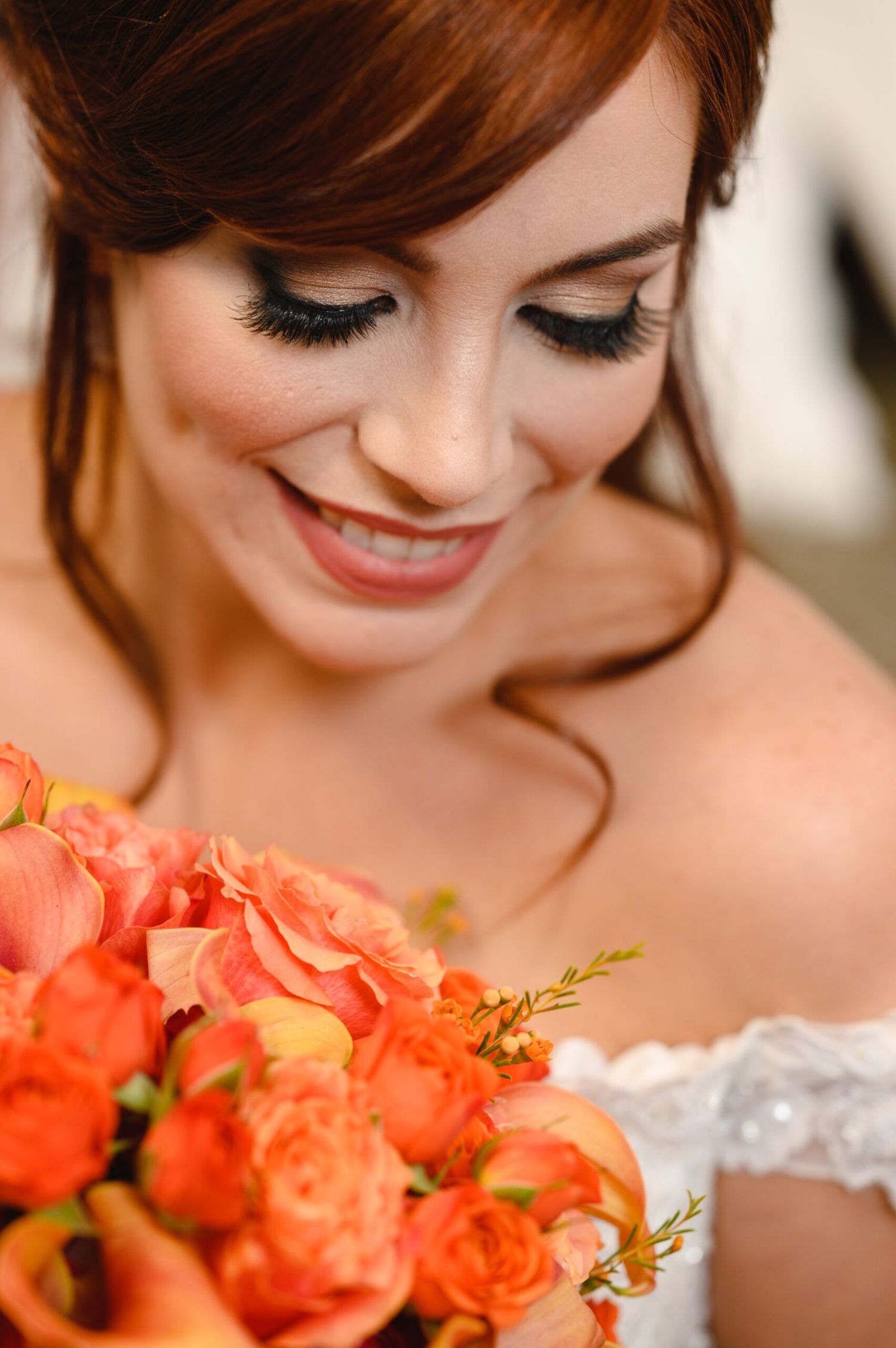 Smiling redhead bride with dramatic lashes holds vibrant orange and coral flower bouquet in lace wedding dress.