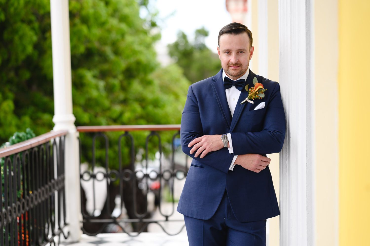 Groom in navy blue suit with boutonniere leans against yellow building on balcony, arms crossed, smiling.