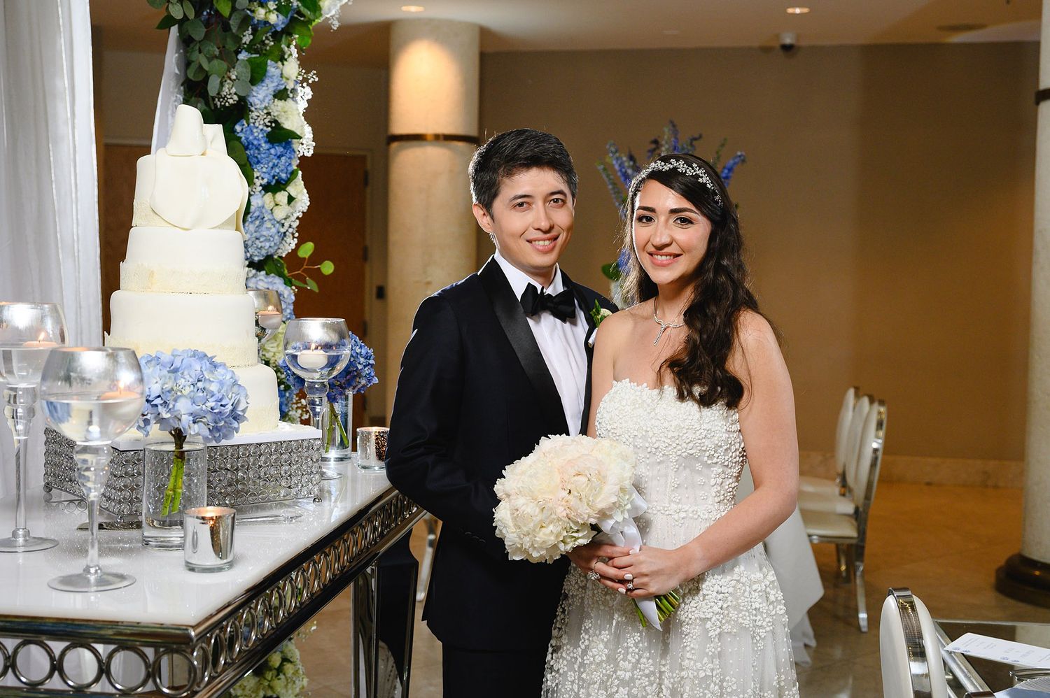Bride and groom smiling together at wedding reception beside elegant white tiered cake with blue floral arrangements.