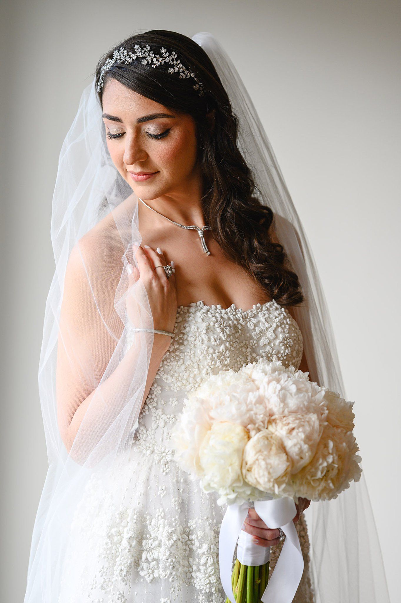 Bride in beaded strapless gown with veil and crystal headpiece holds white peony bouquet, looking down gracefully.