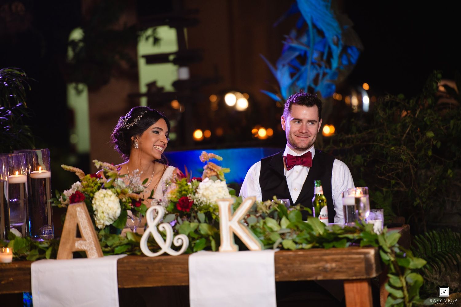 Bride and groom smiling at sweetheart table decorated with floral centerpiece and monogram letters A and K at wedding reception.