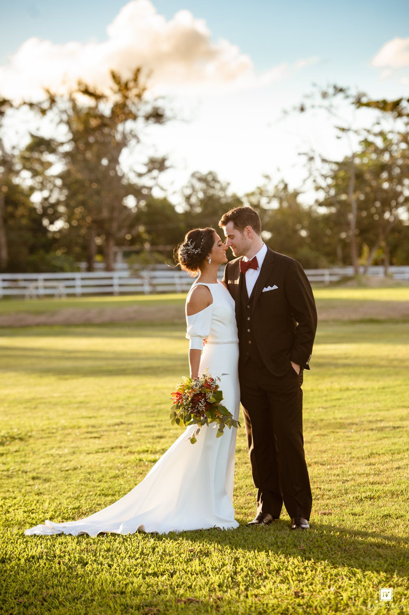 Bride and groom share a romantic moment on lush green lawn, she in white gown with bouquet, he in black tuxedo with red bow tie.