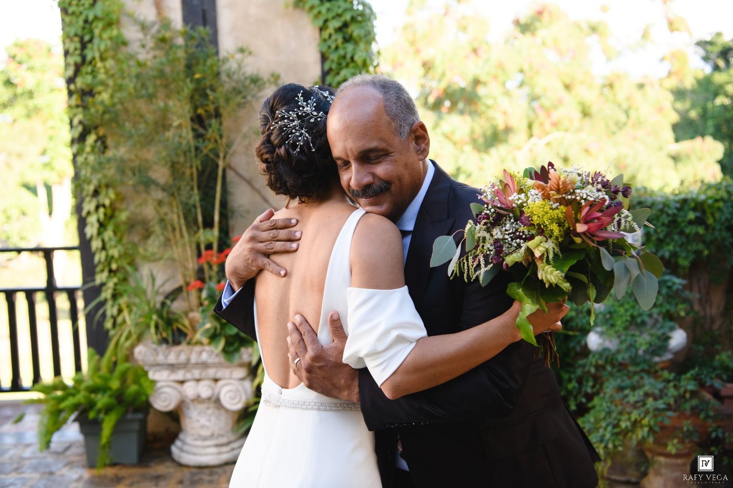 Bride in white gown receives heartfelt hug from older man holding colorful bouquet at outdoor wedding venue.