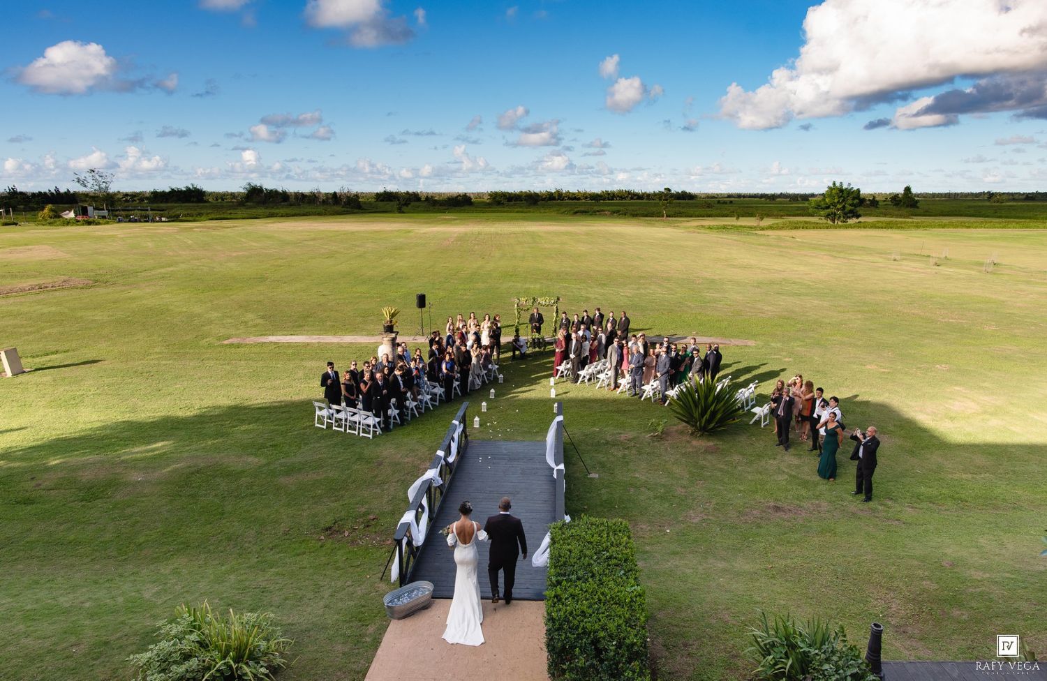 Aerial view of an outdoor wedding ceremony on a lush green lawn with guests seated in a semicircle under a blue sky.