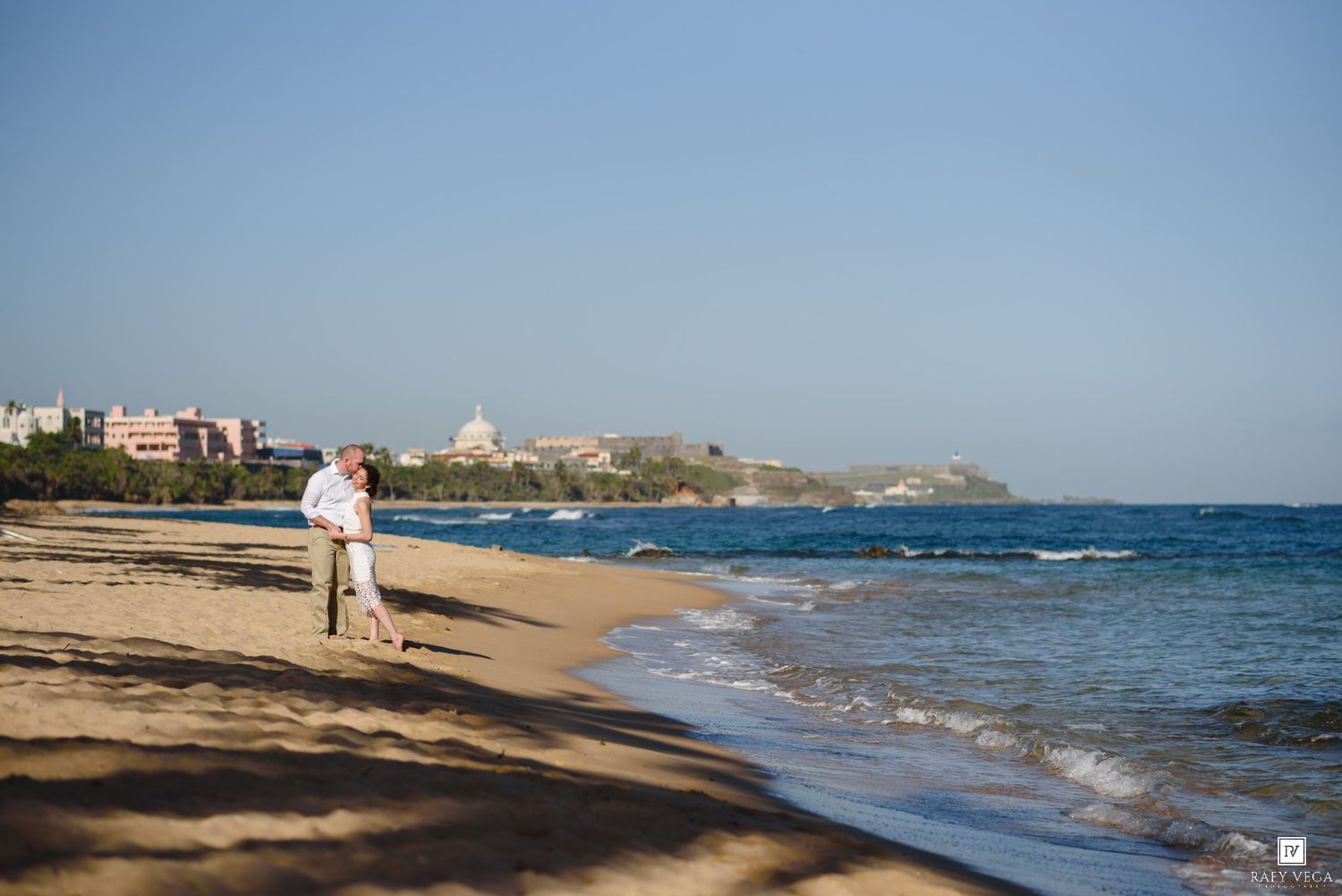 Couple walking hand in hand on a sandy beach with a coastal city skyline in the background.