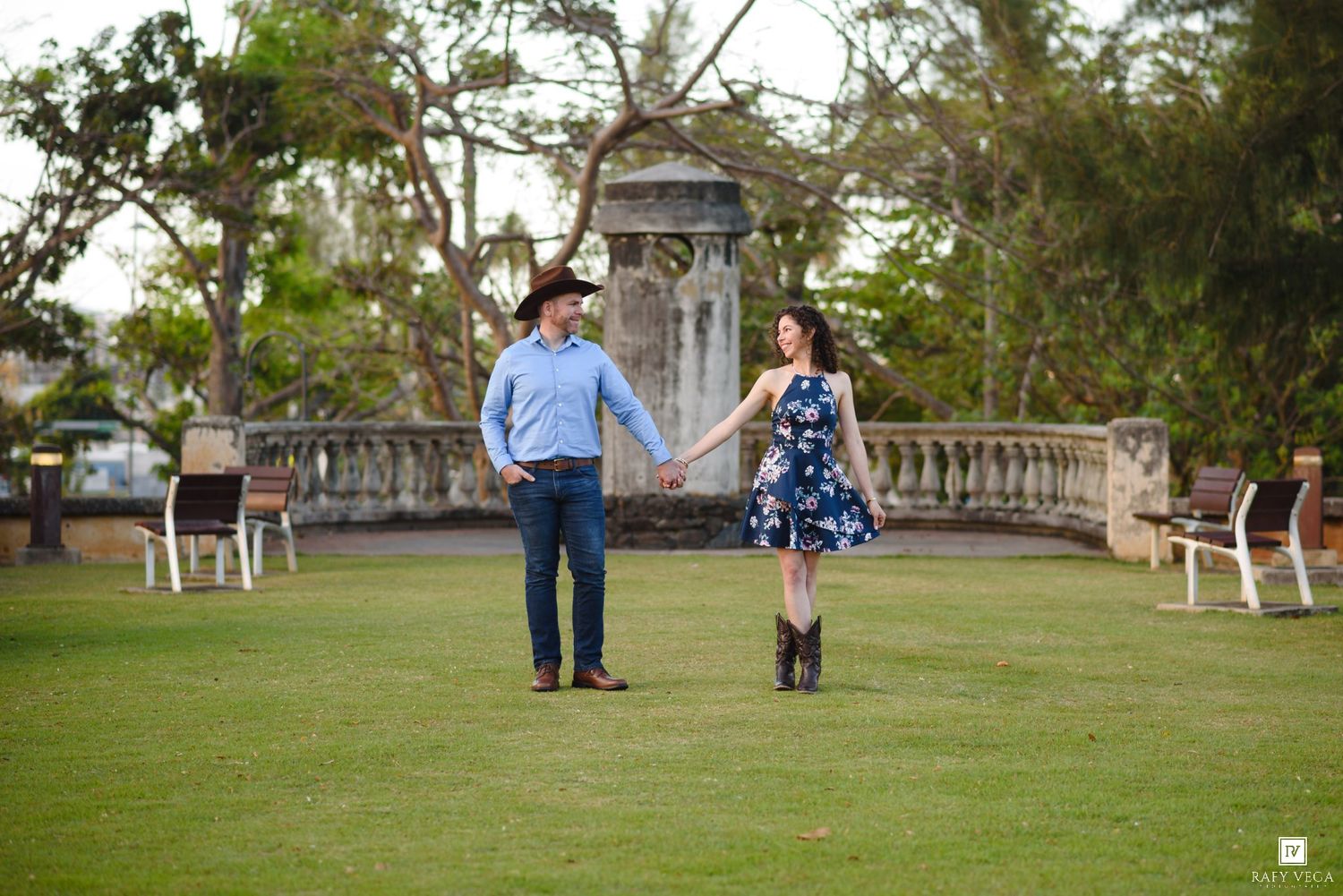 Couple holding hands on green lawn with stone balustrade and trees in background during outdoor engagement photo session.