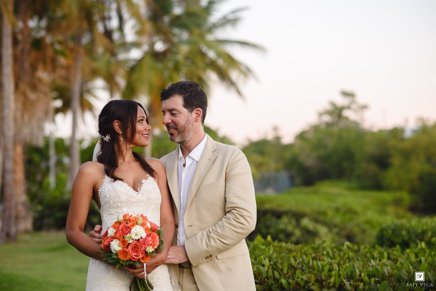 Bride and groom share a romantic moment outdoors with tropical palm trees and lush greenery in a destination wedding setting.