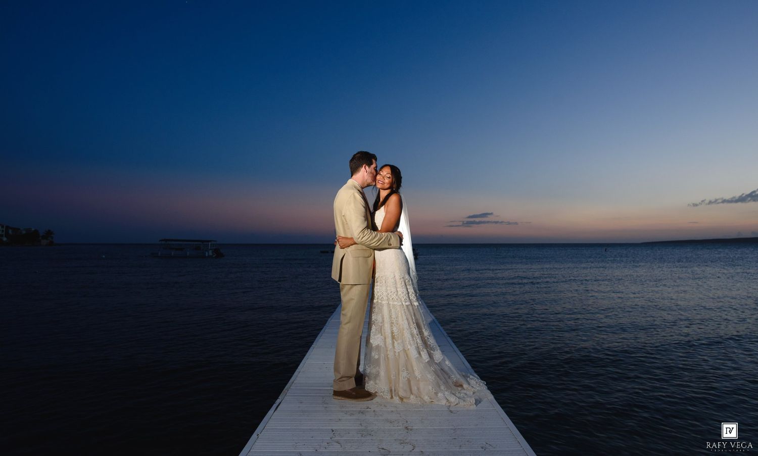 Couple sharing a romantic kiss on a pier at sunset over calm ocean waters during their beach wedding.