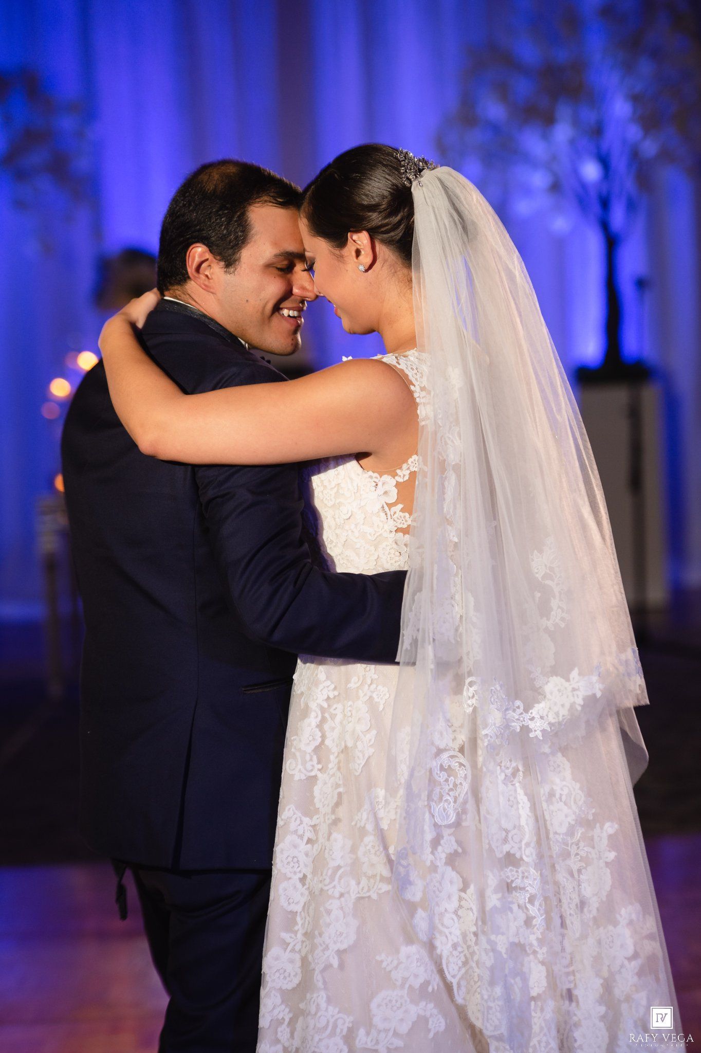 Newlyweds embrace during first dance, wearing lace wedding gown and navy tuxedo in elegant blue-lit venue.