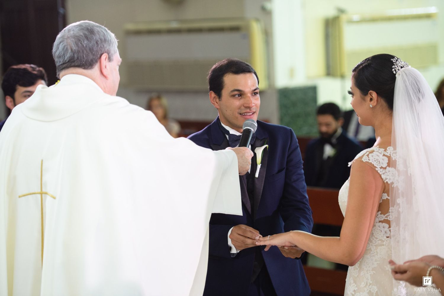 Groom smiling at bride during wedding ceremony while priest officiates in a church.