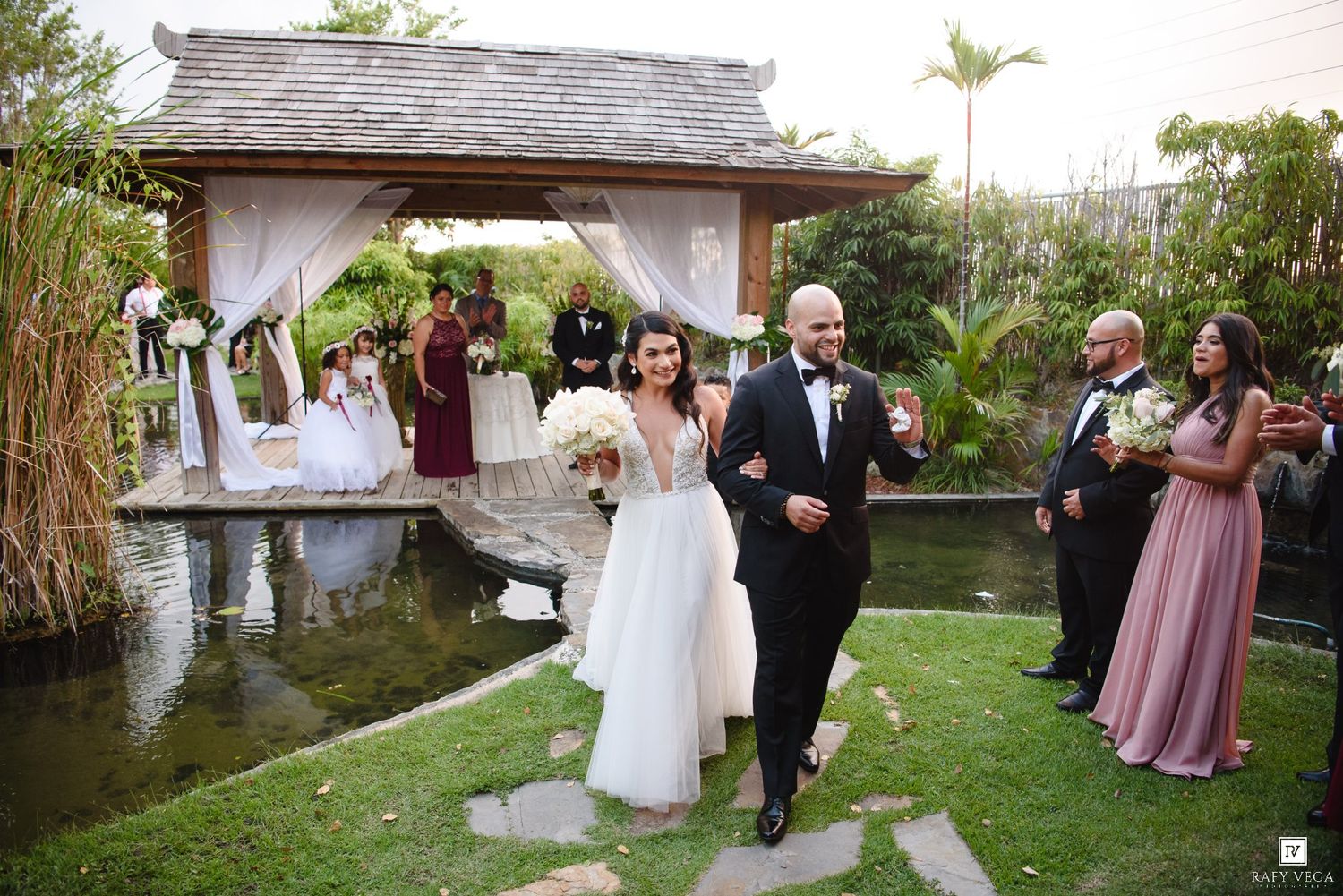 Newlyweds walk down garden path after outdoor wedding ceremony with guests celebrating.