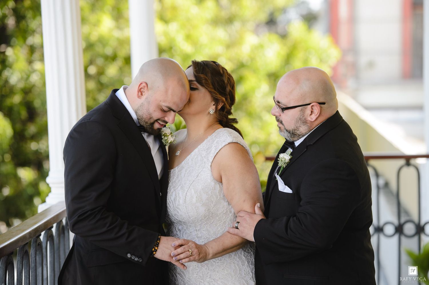 Bride kissing groom while officiant stands beside them on a decorative balcony during an outdoor wedding ceremony.