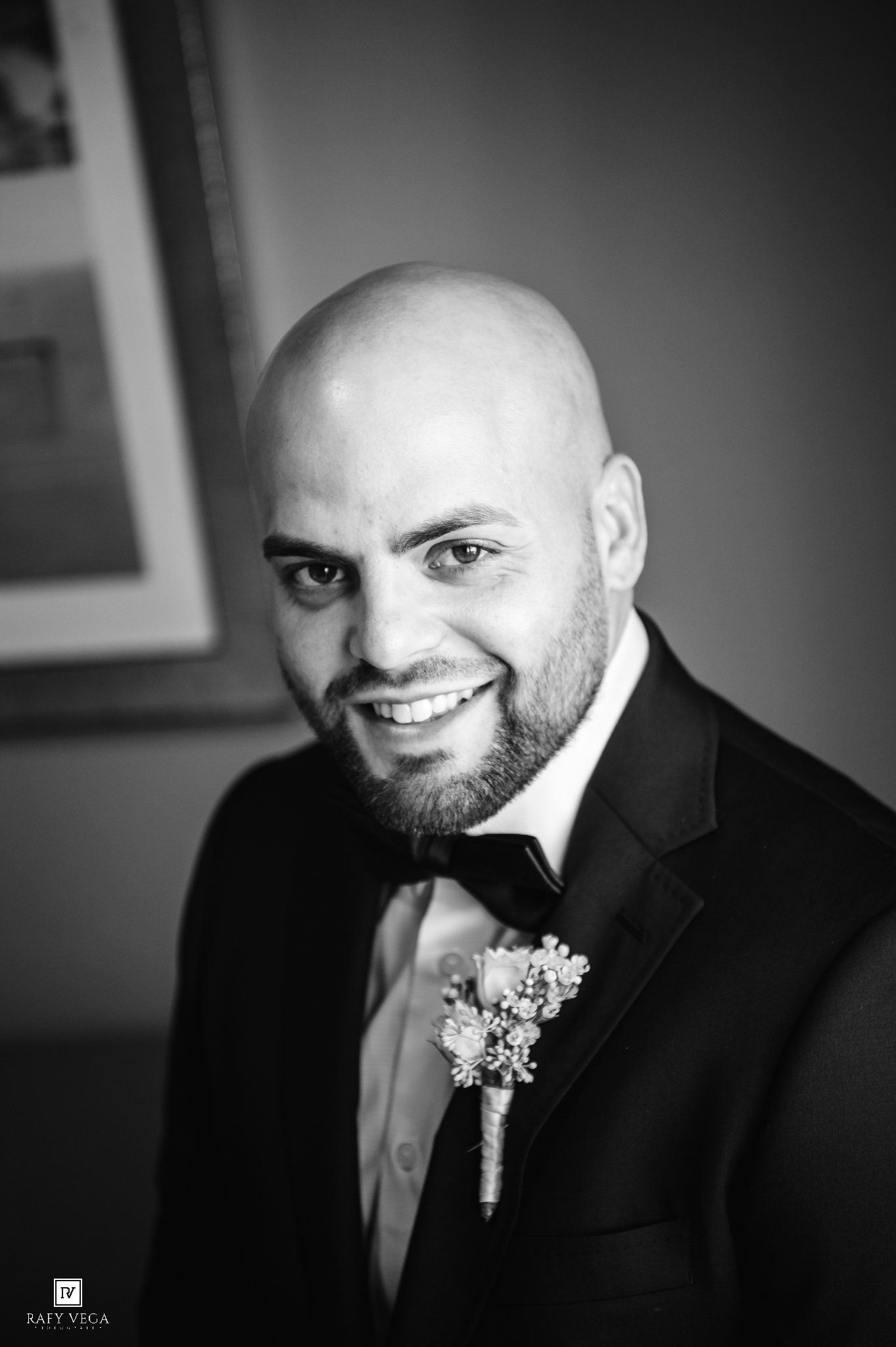 Smiling bald groom with beard wearing tuxedo and boutonniere in black and white wedding portrait photo.