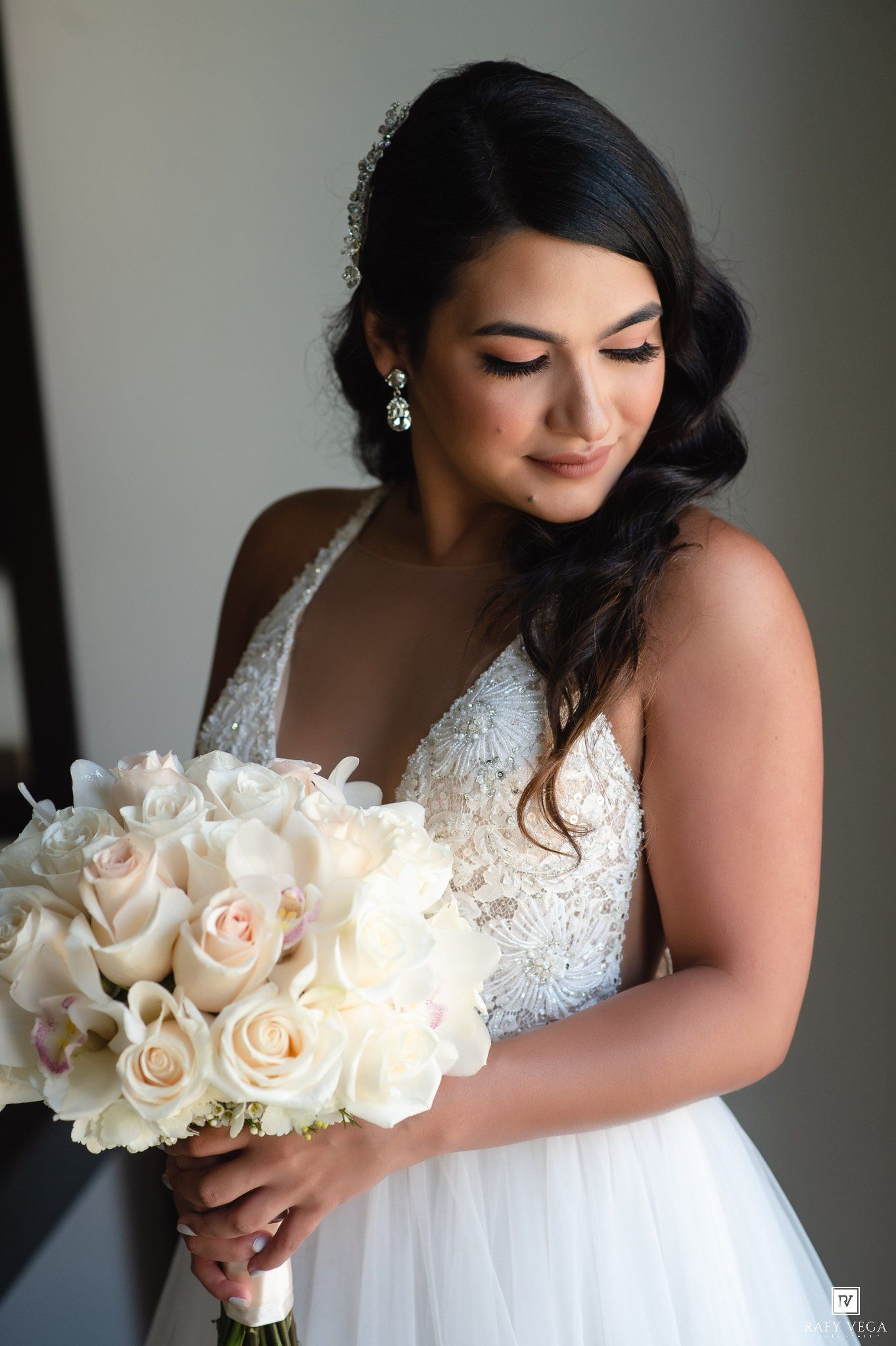 Bride in white lace ballgown holding ivory rose bouquet, looking down with elegant side-swept hair and crystal earrings.