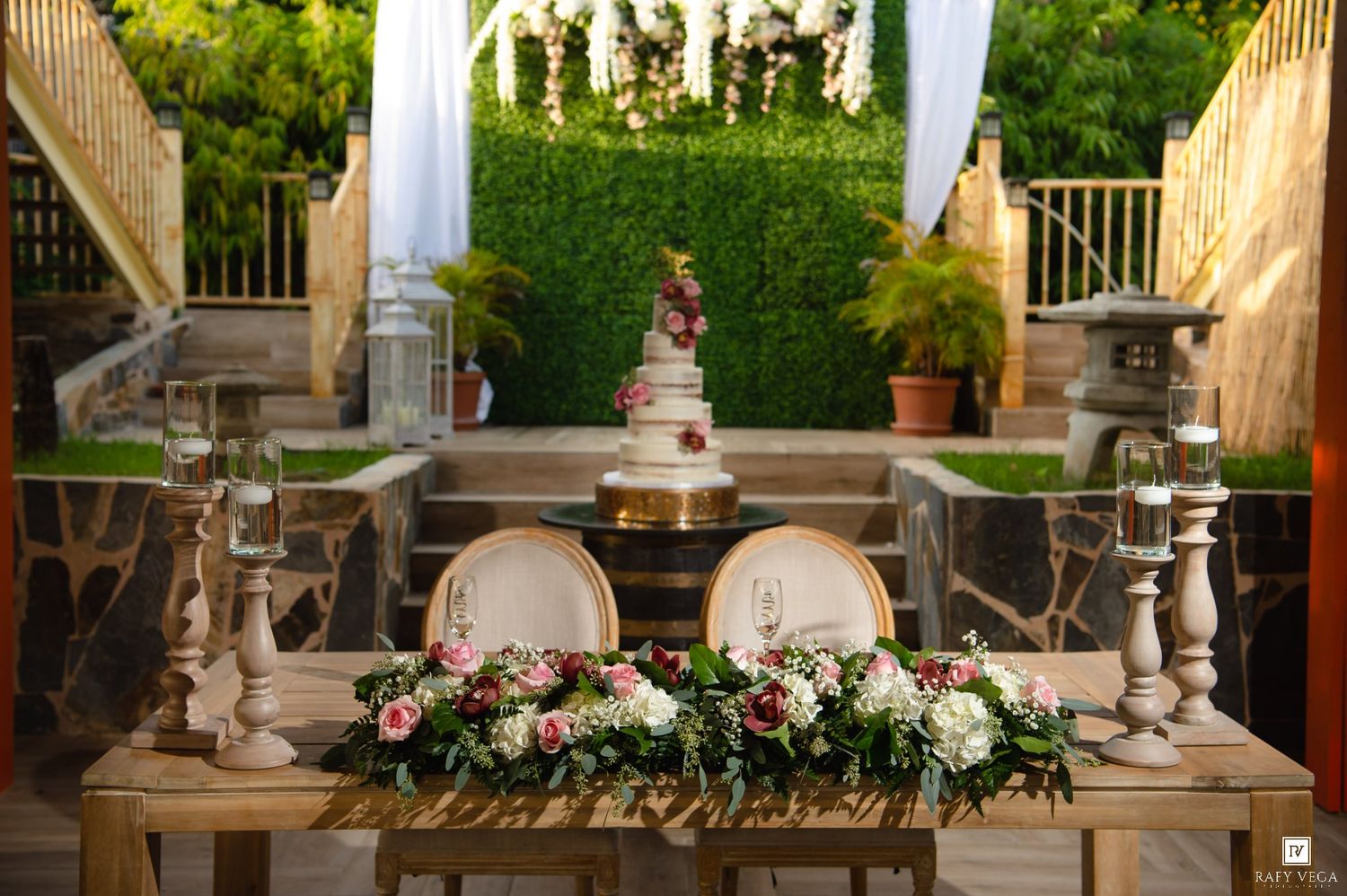 Outdoor wedding cake table adorned with white floral arrangements, candle holders, and lush green hedge backdrop.