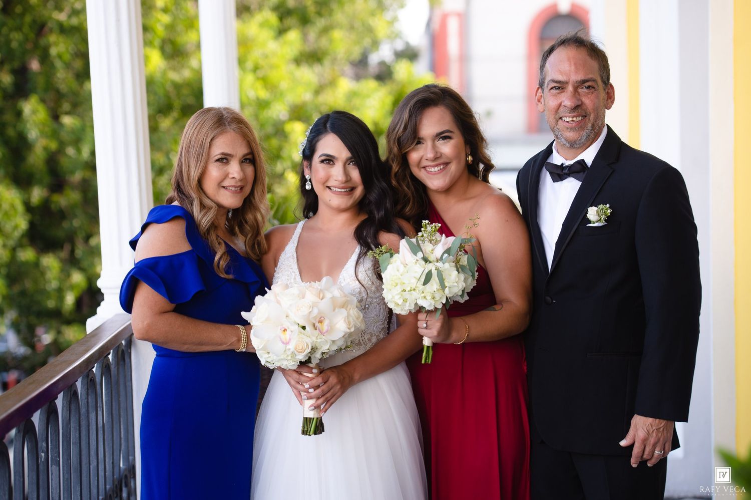 Bride in white gown poses with family members in formal attire on a porch, holding white bouquets.
