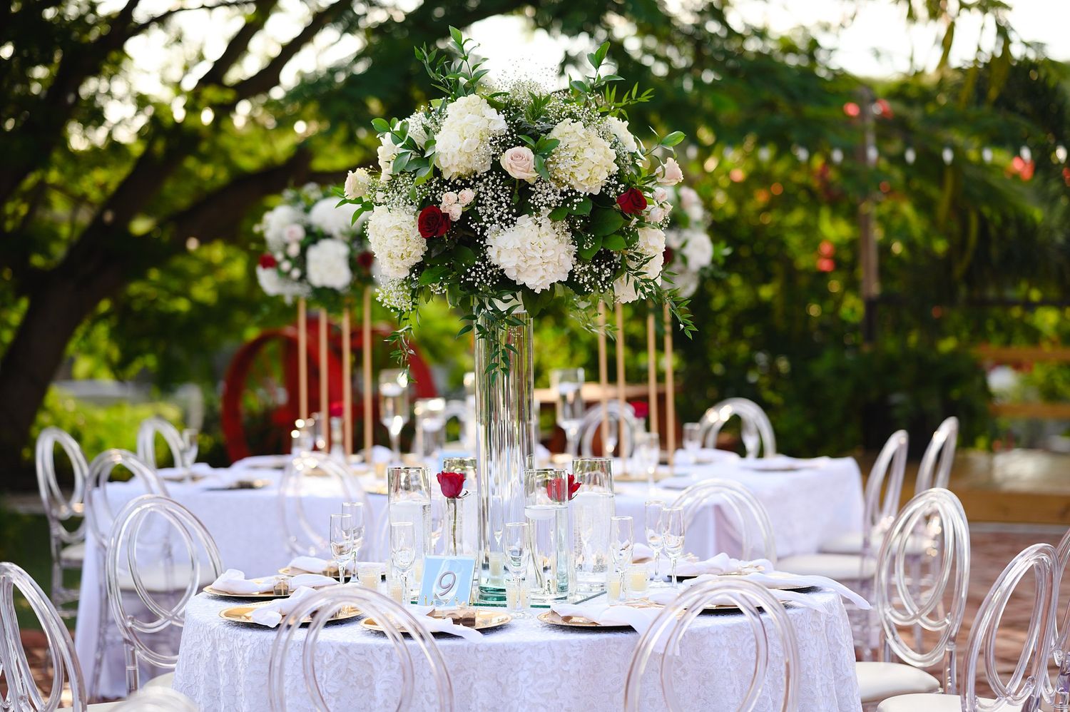 Elegant outdoor wedding reception table with white floral centerpiece, crystal glasses, and ghost chairs under lush green trees.
