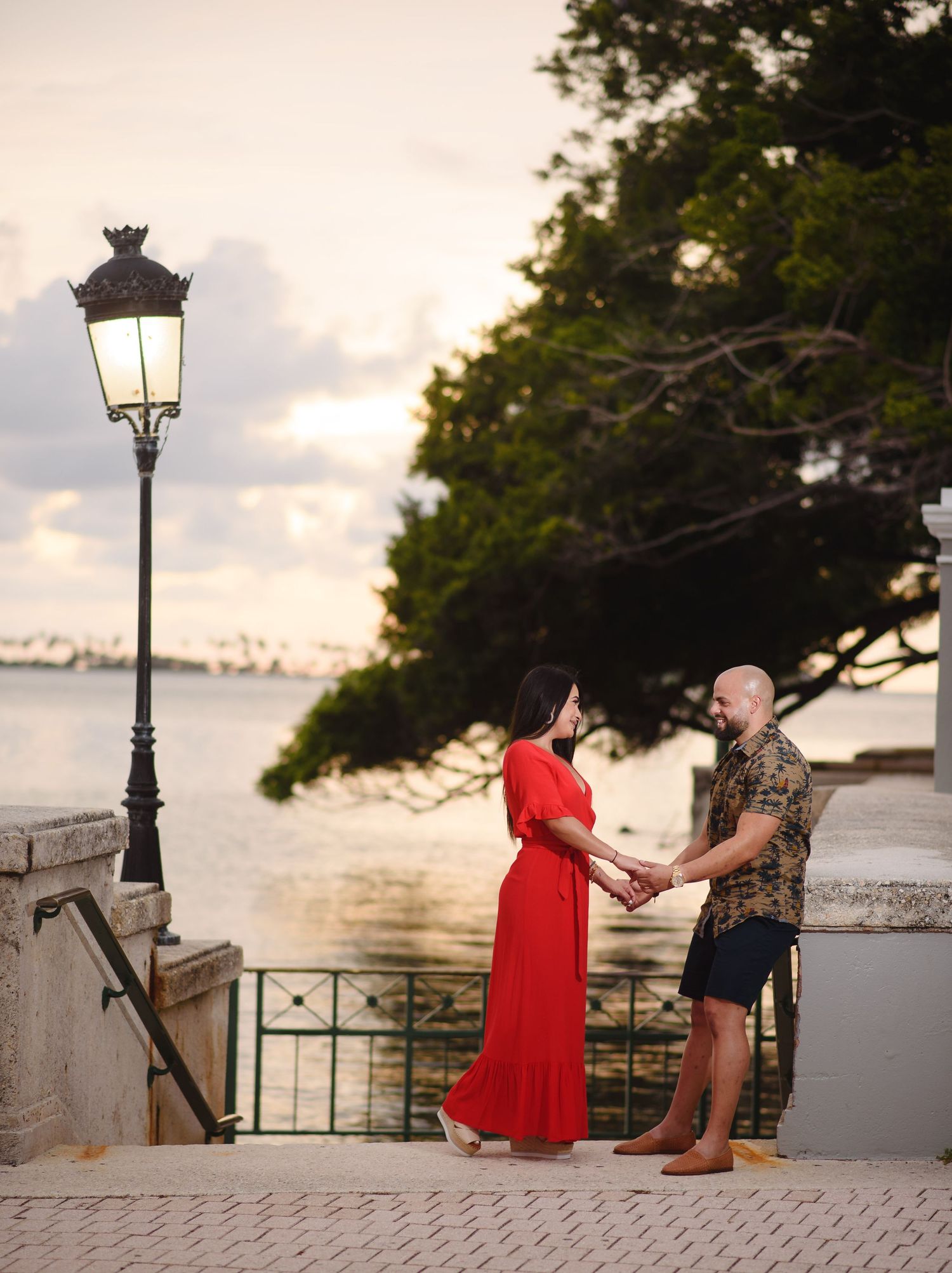 Couple holding hands in red dress and floral shirt at sunset by waterfront with vintage lamp post.
