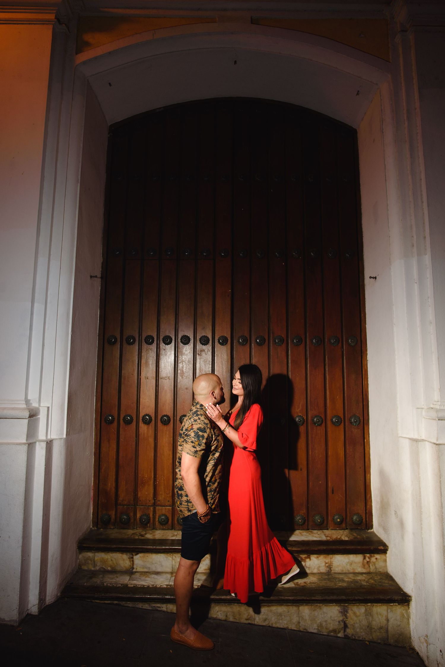 Couple in romantic embrace at night, woman in red dress and man in floral shirt, standing before a grand wooden door.