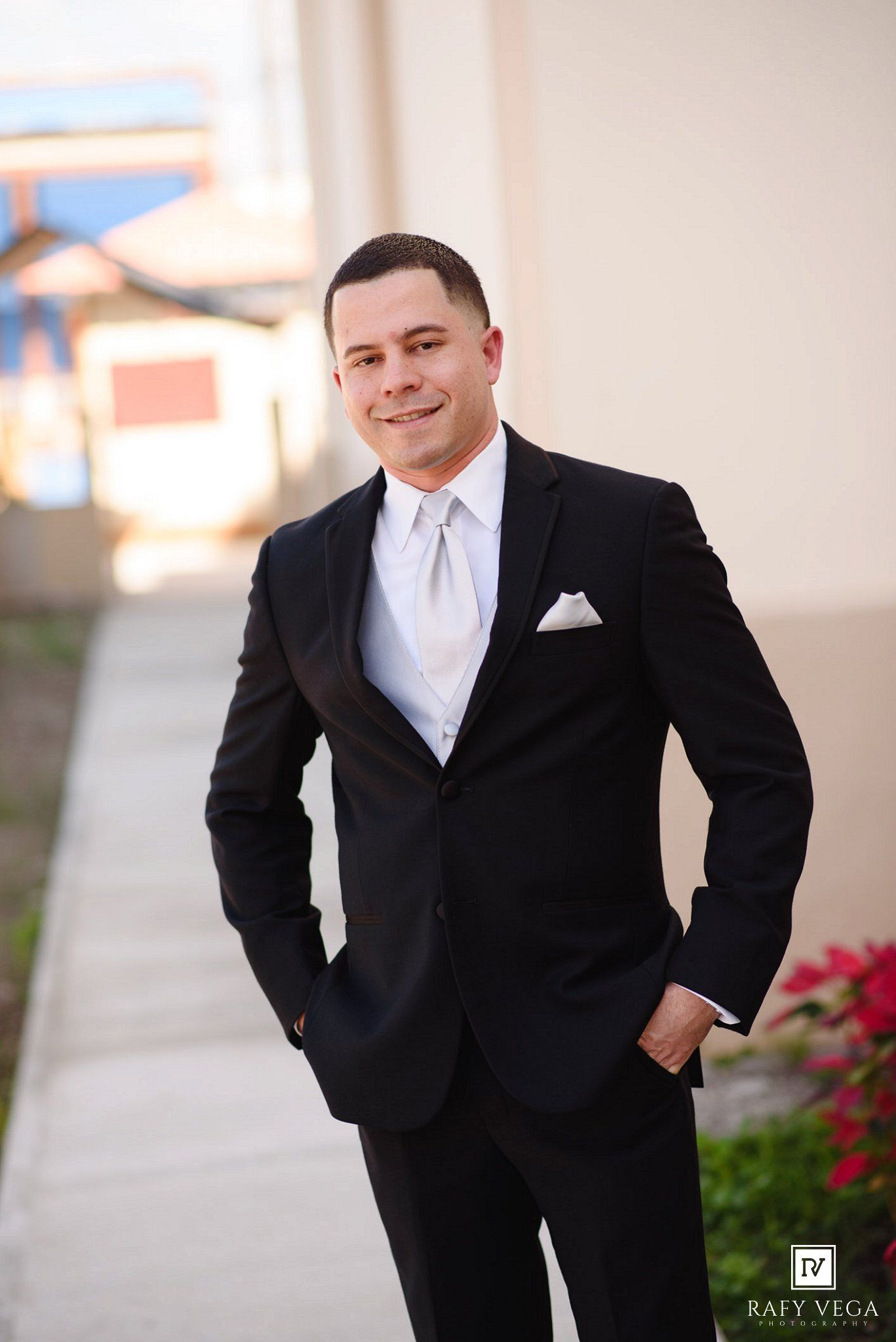 Groom in black tuxedo with white vest and tie posing outdoors.