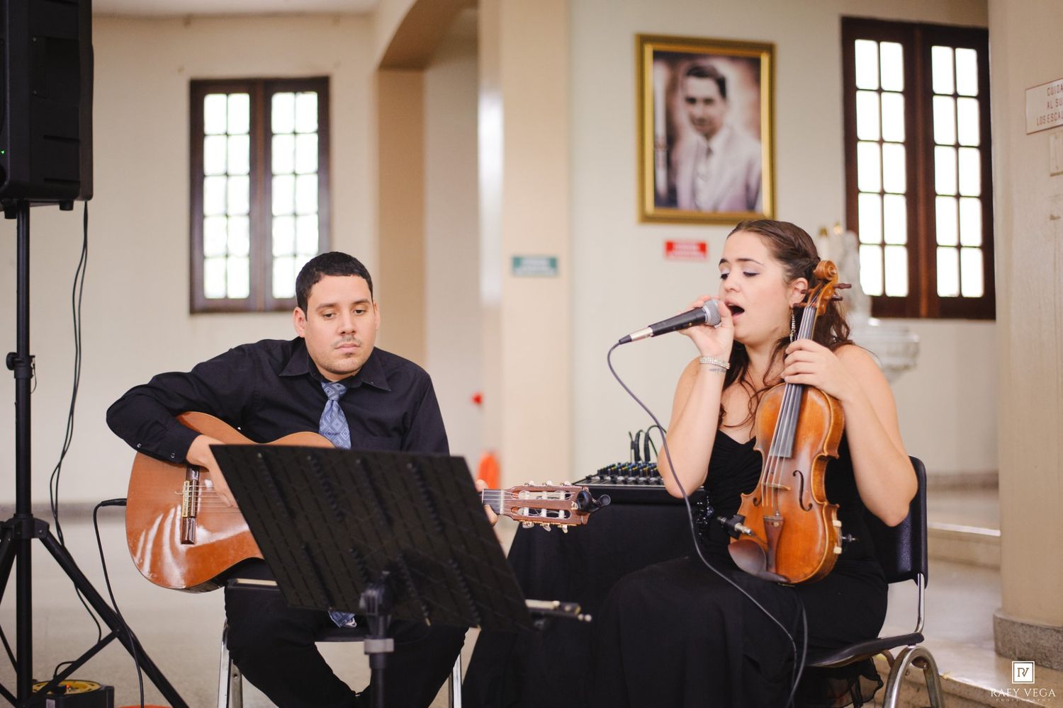 A man playing acoustic guitar and a woman singing into a mic while holding a violin perform together indoors.