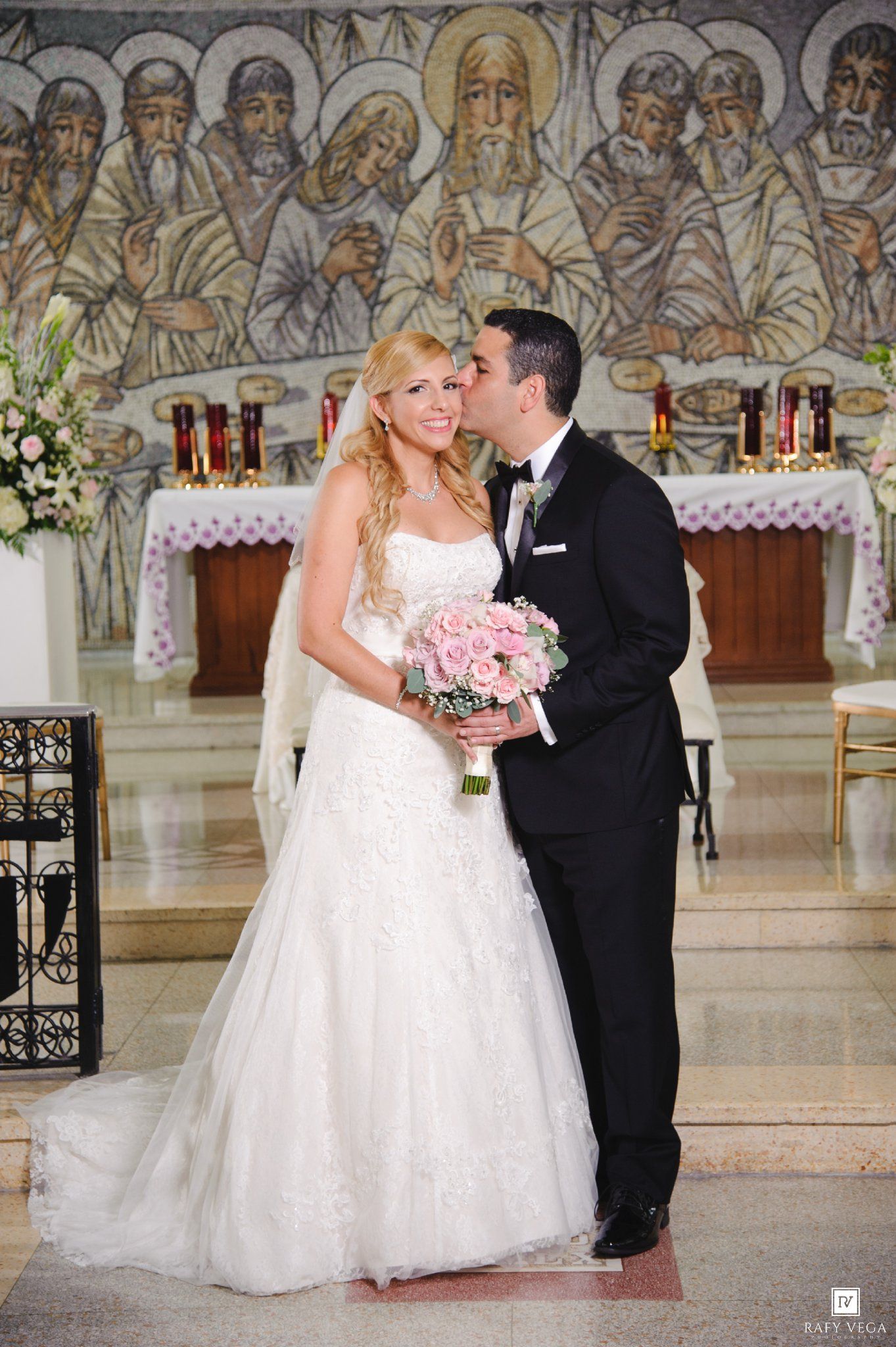 Groom kisses bride on cheek as she holds pink rose bouquet in front of ornate church mosaic altar.