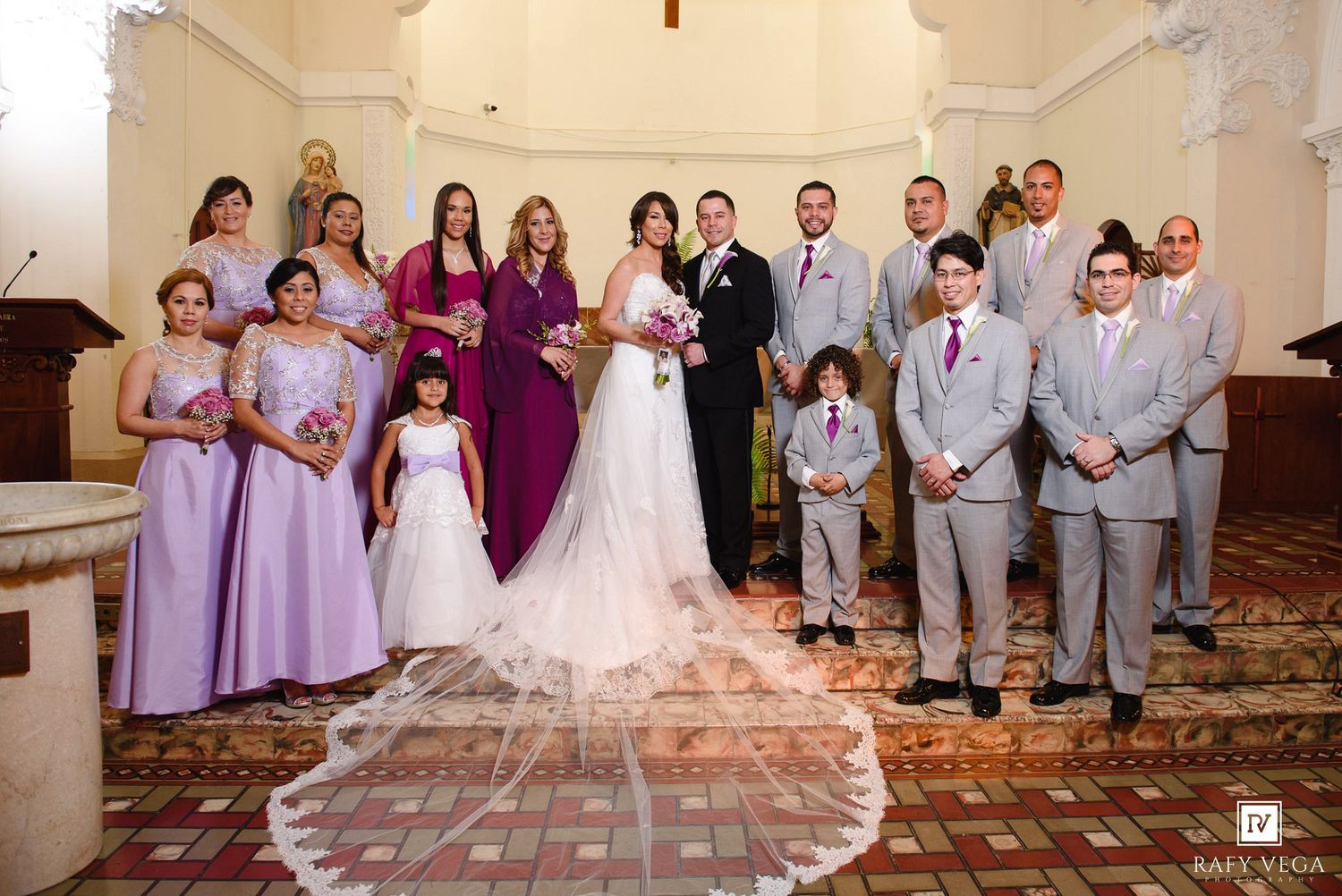 Wedding party poses in church with bride in white gown, bridesmaids in purple dresses, and groomsmen in gray suits.