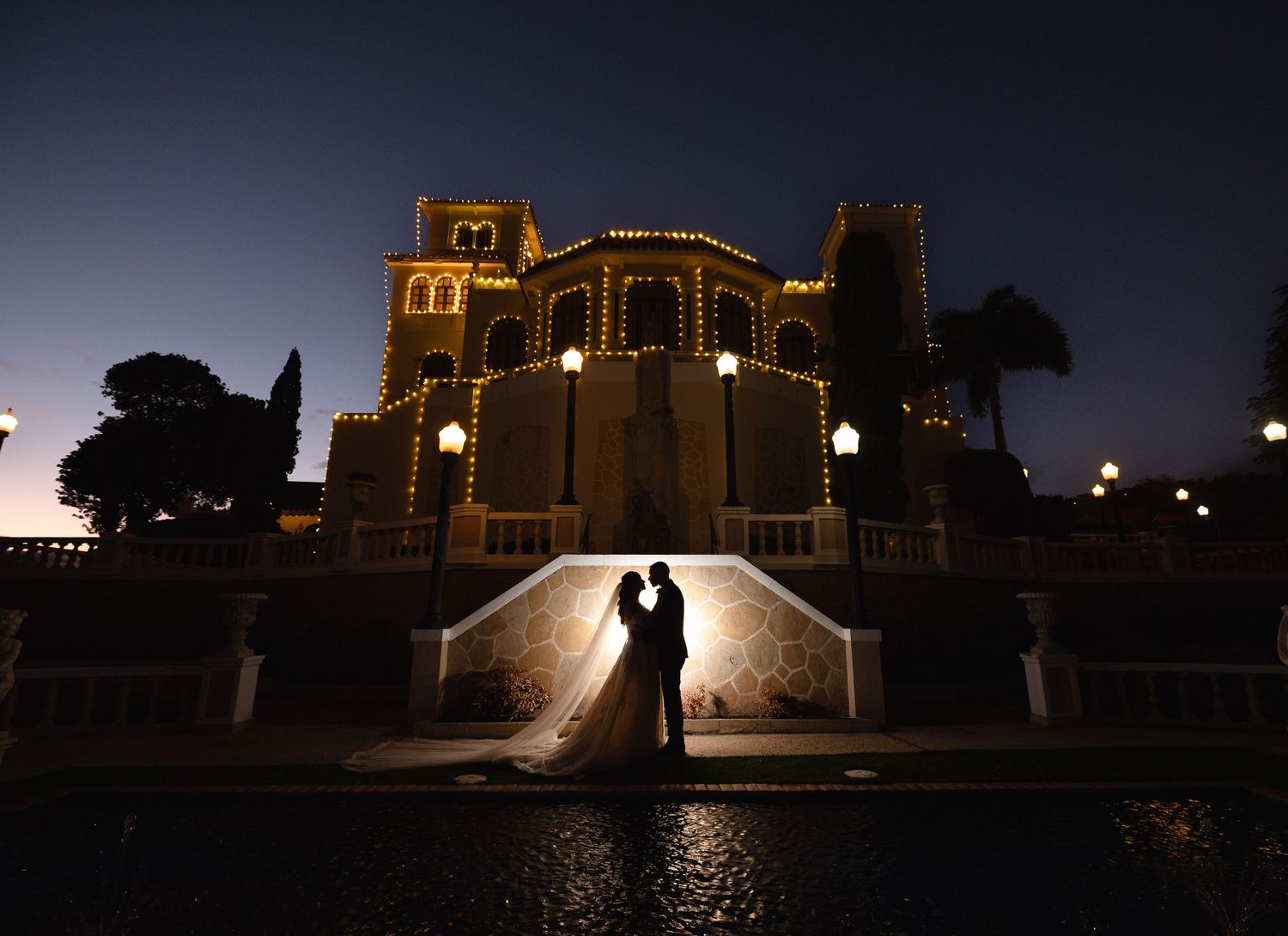 Romantic wedding couple silhouetted against an illuminated historic building at dusk.